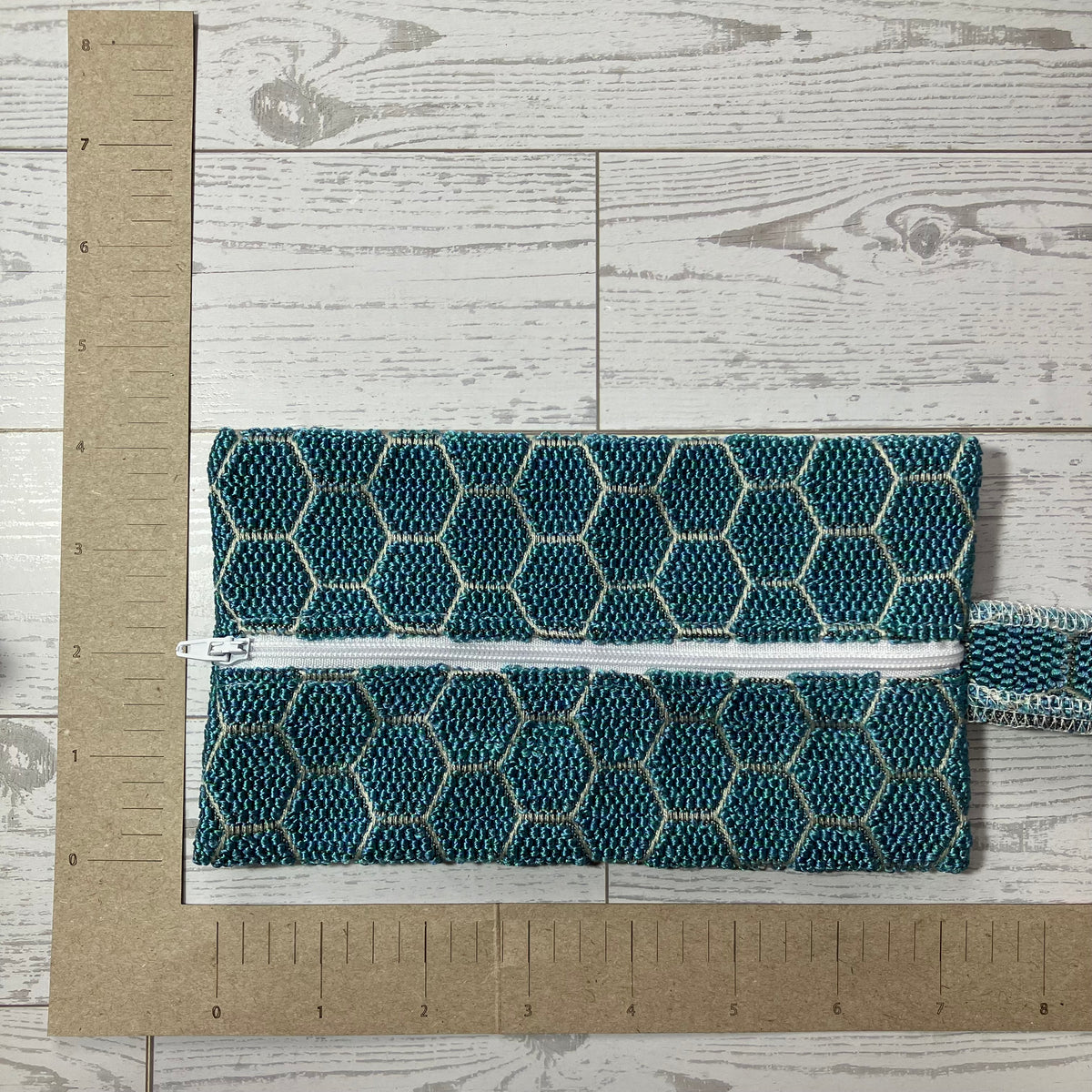 A teal and white honeycomb-patterned zipper pouch with a white zipper, shown on a white wood background with a cardboard ruler for scale.