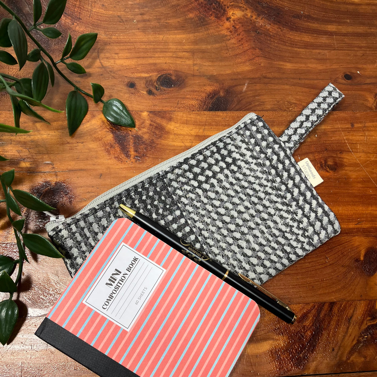 A gray and white patterned fabric zip pouch with a loop handle, styled on a wooden tabletop next to a coral and blue striped notebook and a black pen.