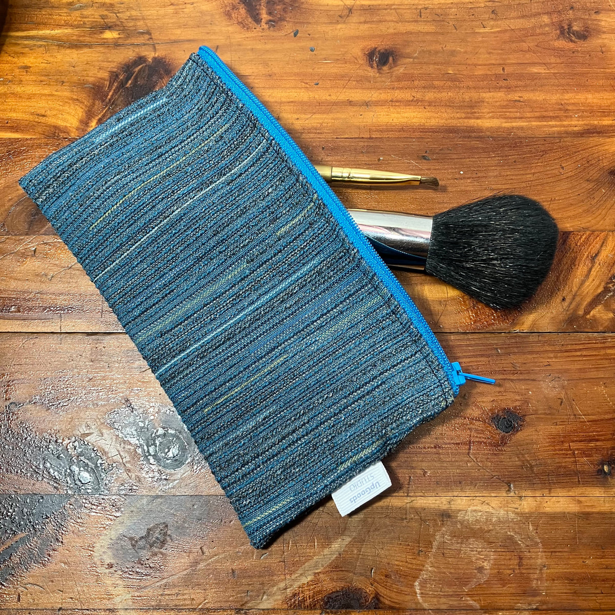 A textured blue makeup pouch with a bright blue zipper, shown on a rustic wooden table with a makeup brush resting behind it.