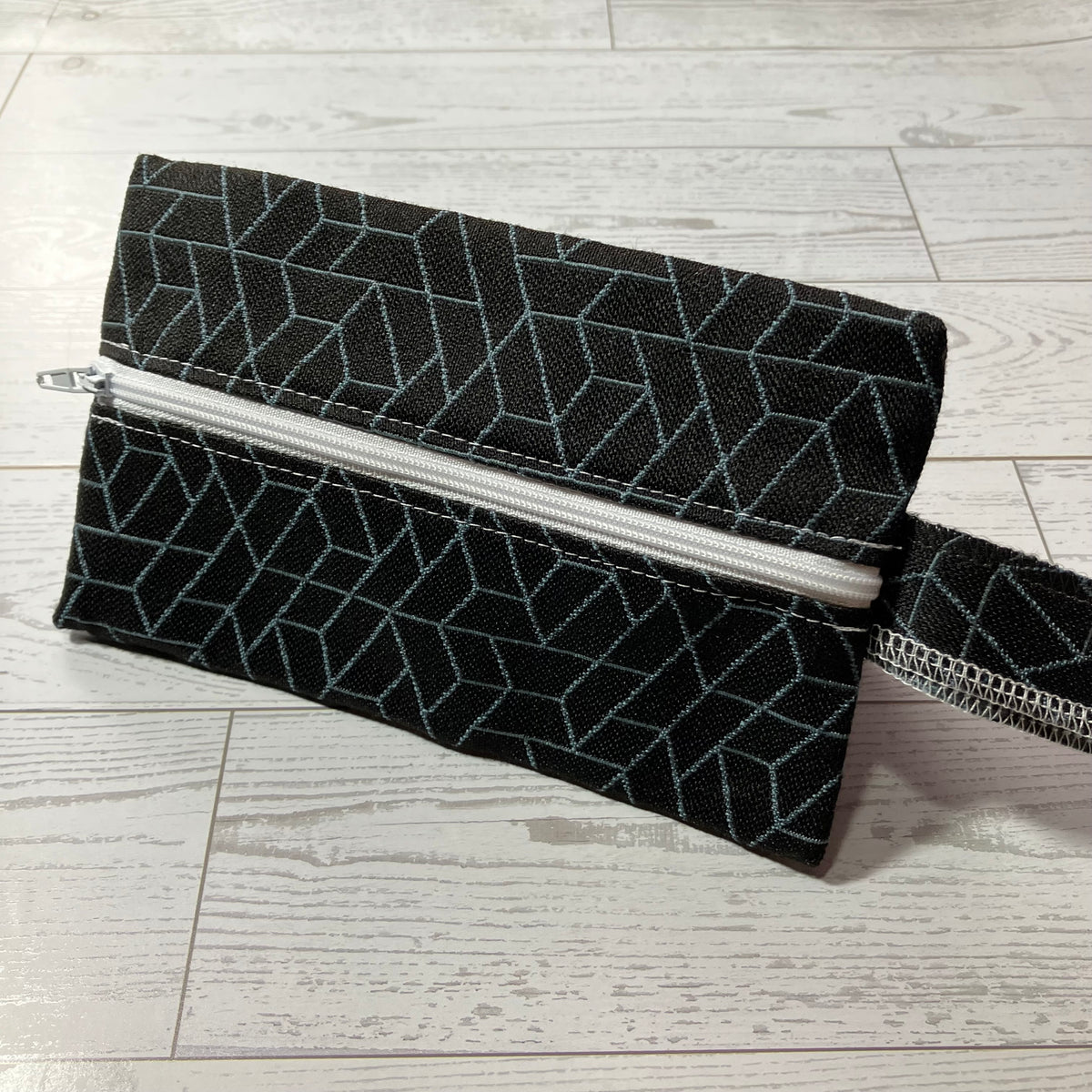 A black fabric zippered pouch with a geometric light blue line pattern, shown on a light gray wood-plank floor.