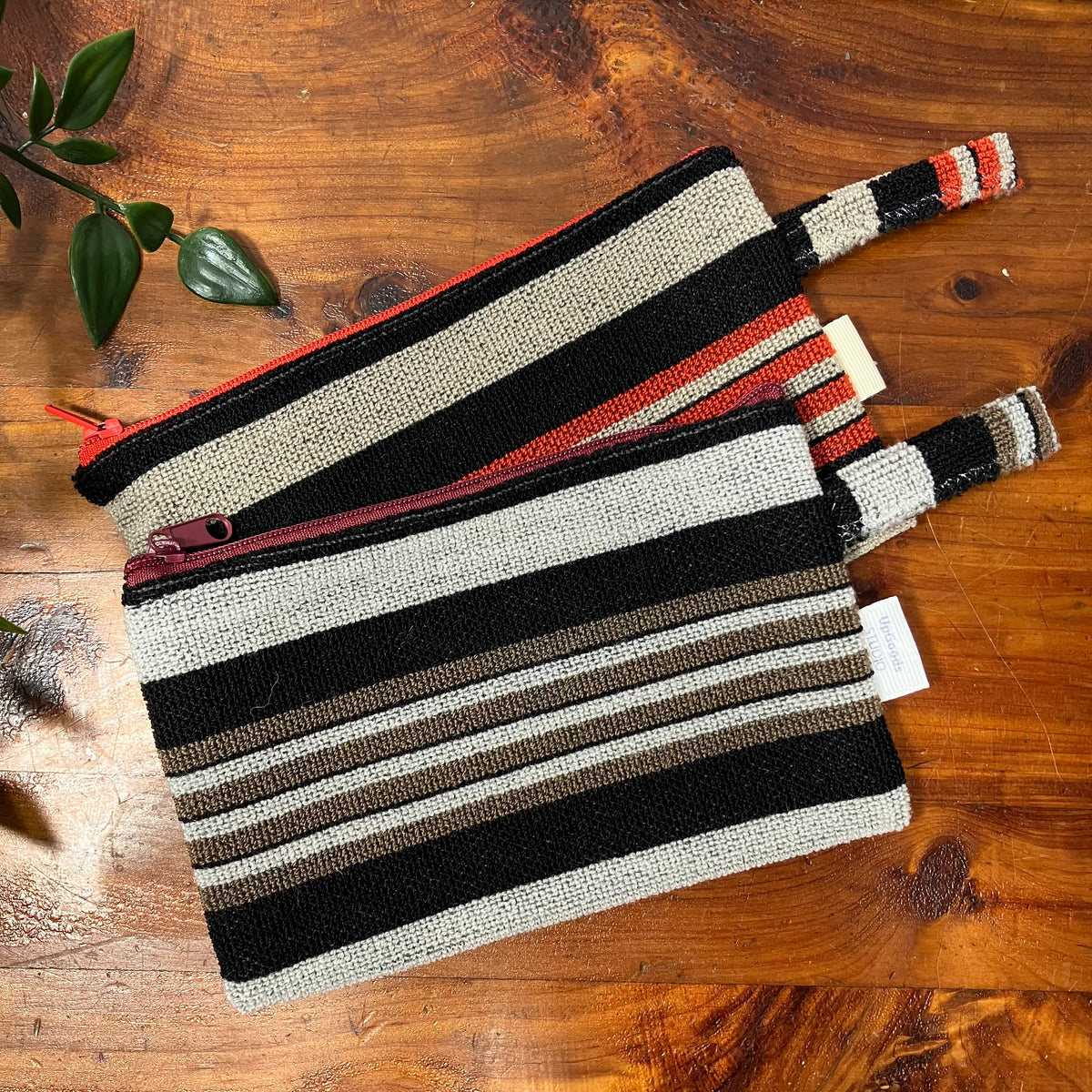Two woven zip pouches with horizontal stripes in black, beige, brown, and red-orange, arranged diagonally on a rustic wooden tabletop.