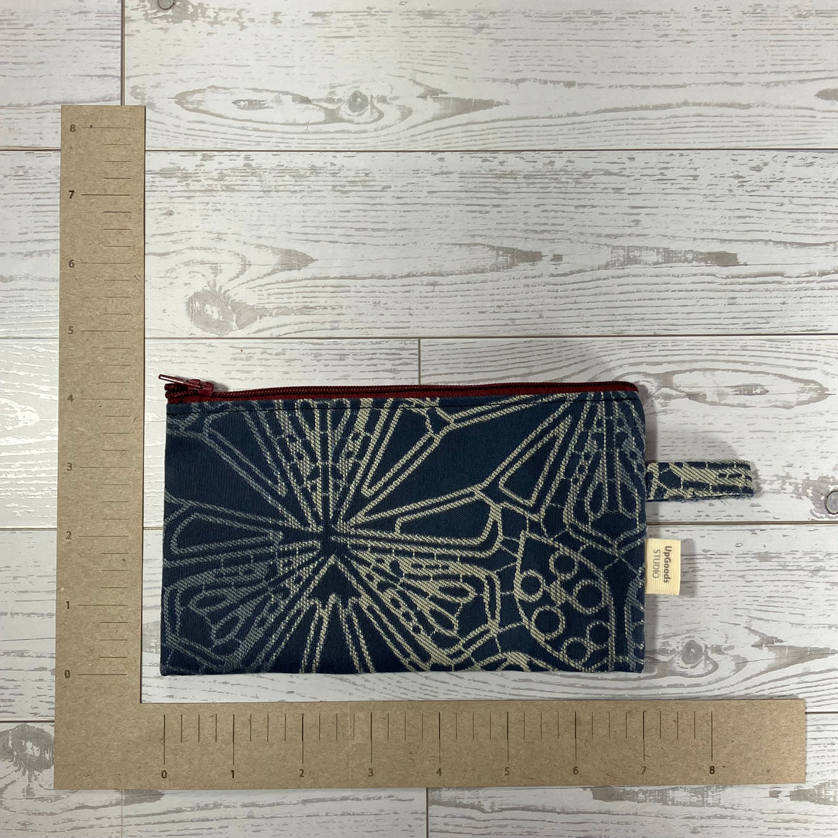 A small fabric pouch or notebook cover with a navy blue and cream geometric pattern, shown flat on a whitewashed wood background with cardboard rulers for scale. A small paper tag reads 'Nanaimo Handmade'.