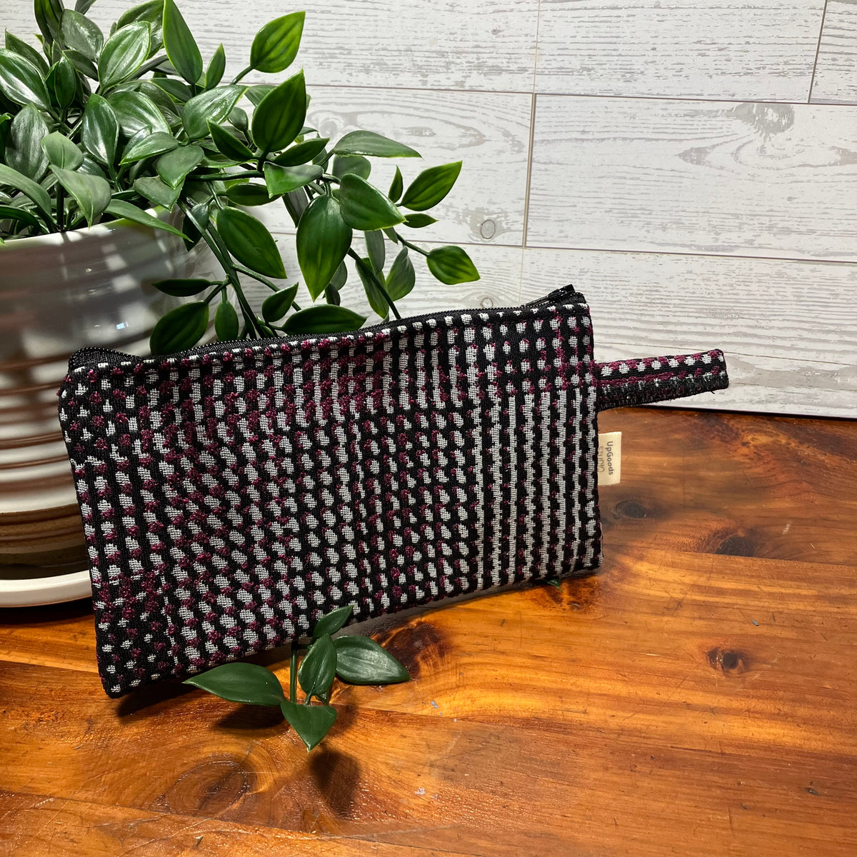 A woven clutch pouch with a small top handle, featuring a black, white, and dark red pattern, resting on a wooden table next to a potted green plant.