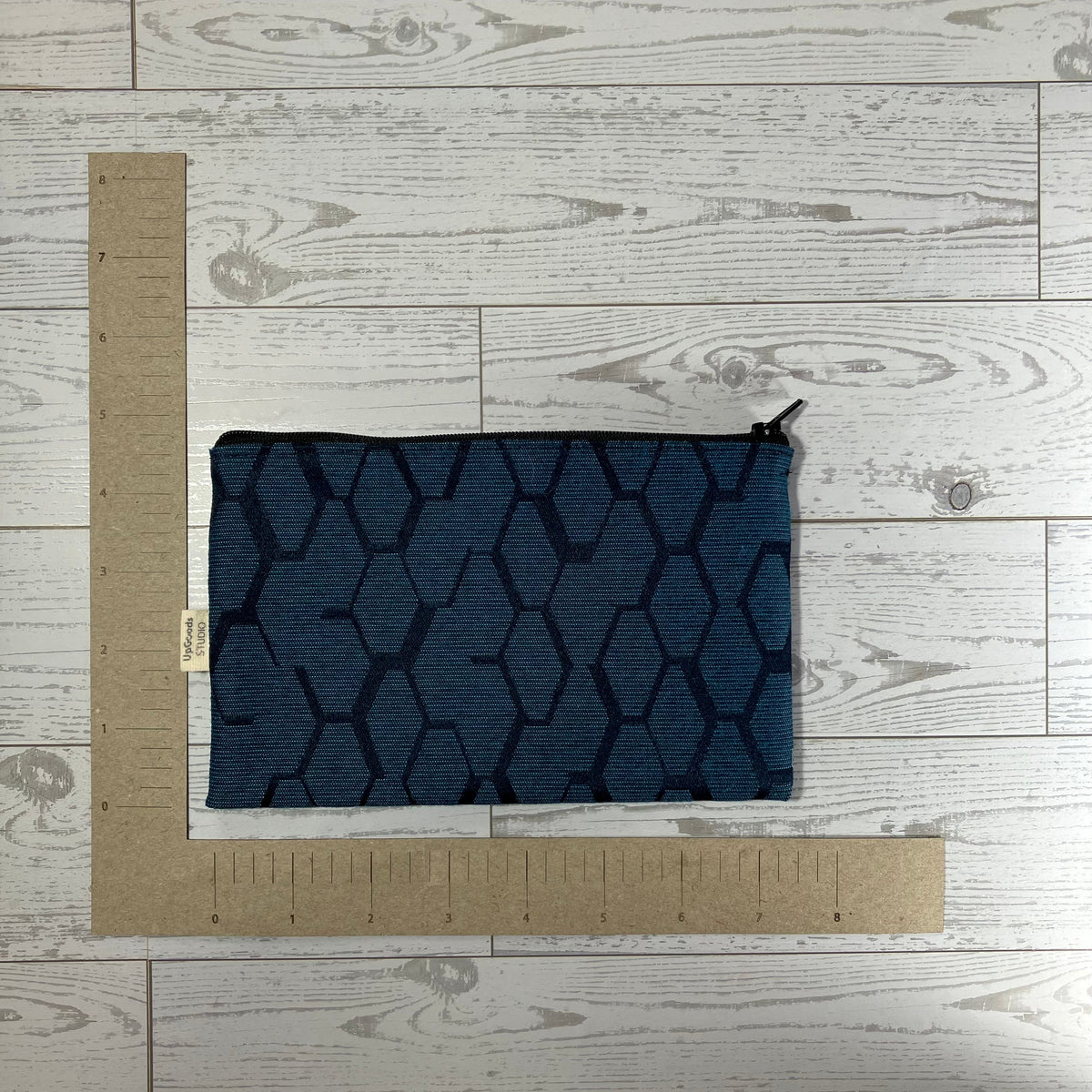 A dark navy blue zippered pouch with a geometric hexagonal pattern, laid flat on a white wood plank background next to a cardboard L-square ruler for scale.