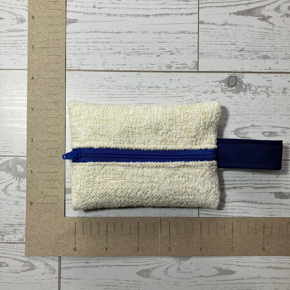 A small woven pouch or organizer in off-white textured fabric with a prominent blue zipper and dark blue fabric tab, photographed on a light wood plank surface with a cardboard ruler for scale.