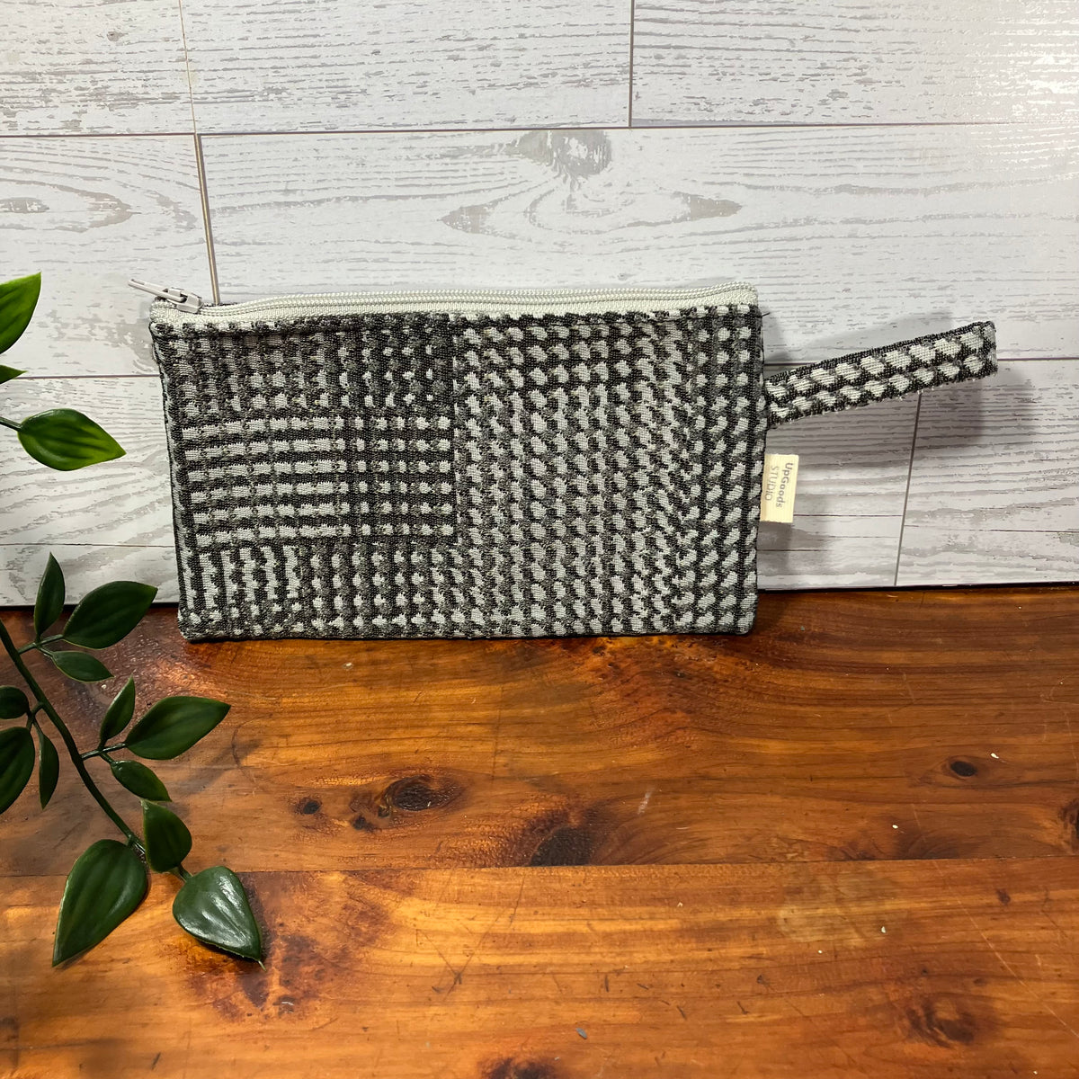 A woven clutch pouch with a black and white houndstooth pattern, resting on a wooden surface against a white wood-paneled wall. A small fabric tag is visible on the right side.
