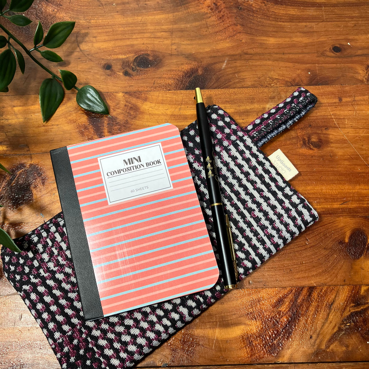 A mini composition book with coral and light blue stripes, a black pen, and a patterned fabric pouch arranged on a rustic wooden tabletop with green leaves in the corner.