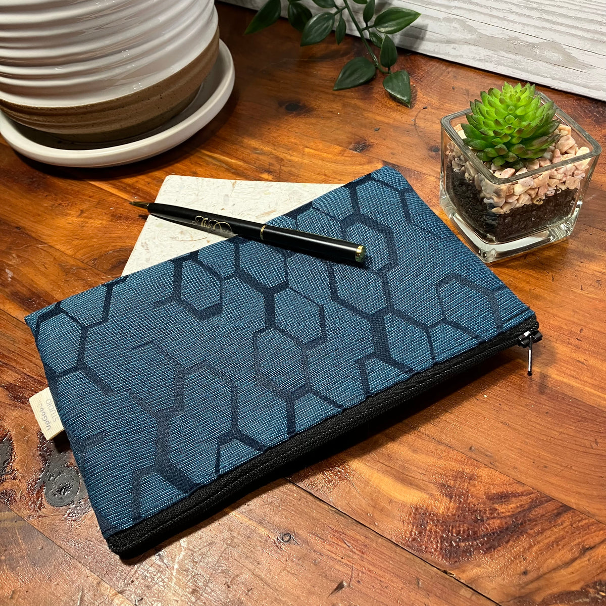 A blue zippered pouch with a black geometric hexagon pattern, styled on a wooden table next to a notebook, a black pen, and a small potted succulent.