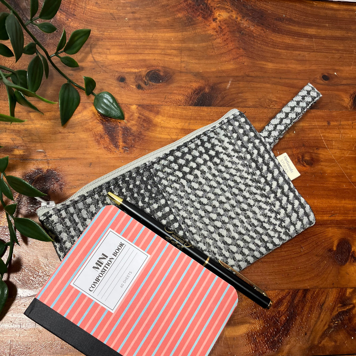A gray and black patterned zippered pouch with a loop handle, styled on a wooden tabletop next to a coral and gray striped notebook and a black pen.