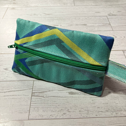 A fabric zippered pouch with a geometric pattern in shades of green, blue, and yellow, resting on a light gray wood plank background.