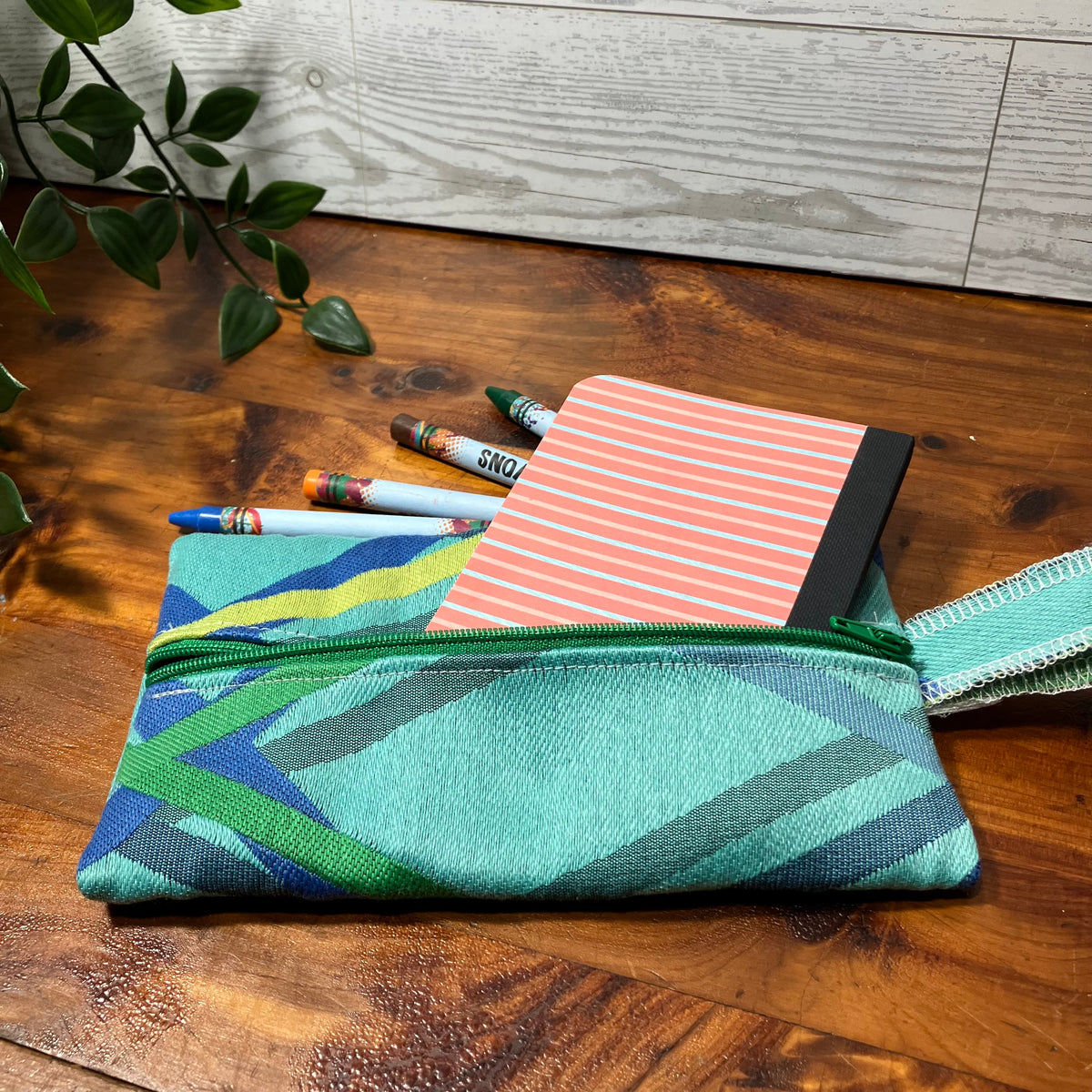 A woven zip pouch with a geometric pattern in teal, green, blue, and gray, shown on a wooden tabletop with a striped notebook and pens inside.