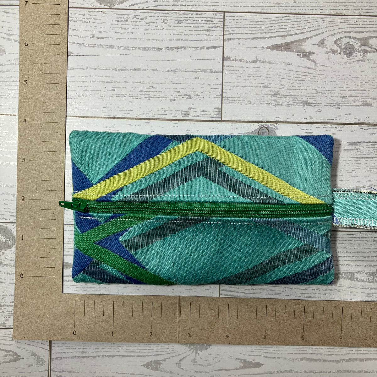 A small zippered fabric pouch with a geometric pattern in teal, blue, green, and yellow, shown on a white wood background with a cardboard ruler for scale.