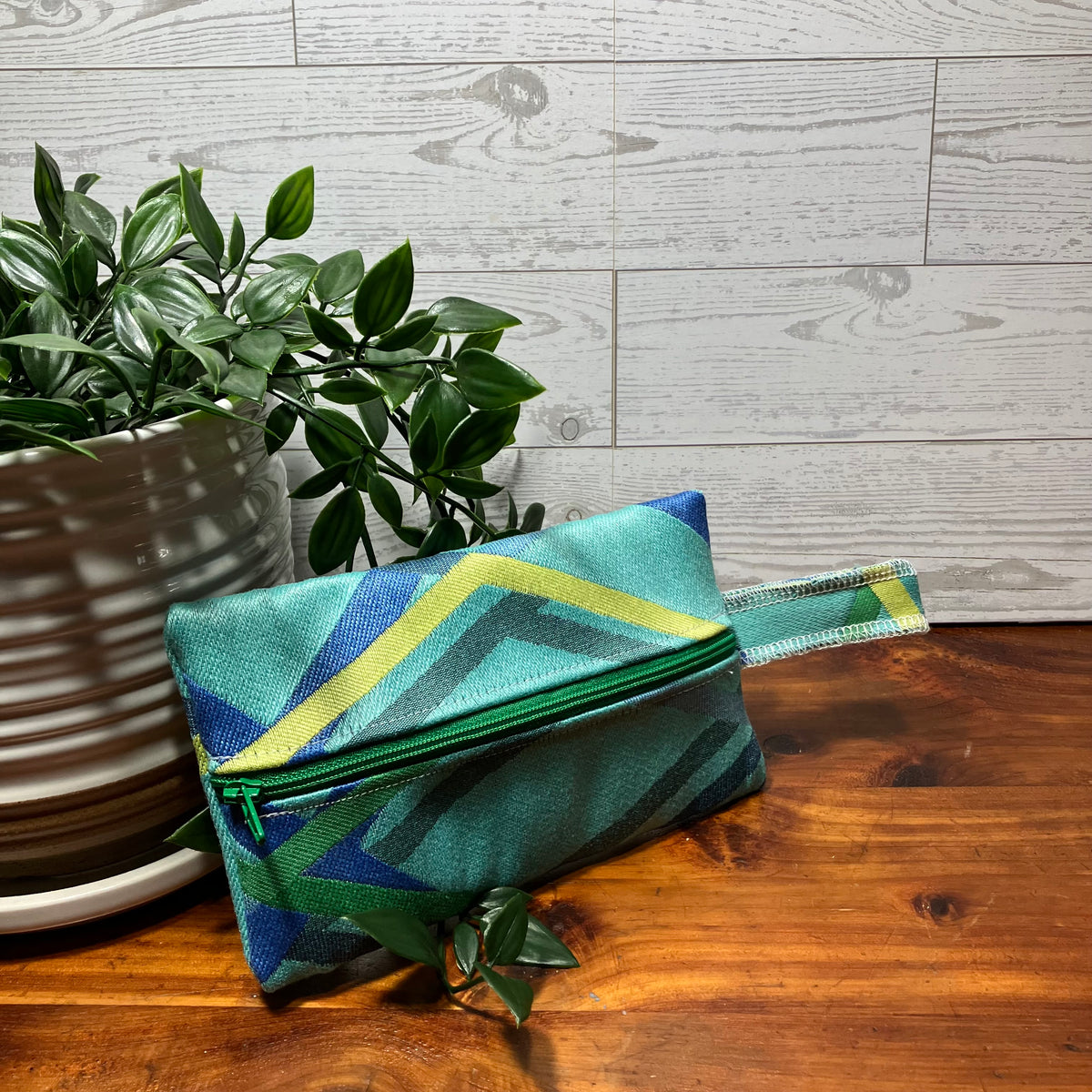 A small zippered pouch with a geometric pattern in teal, green, blue, and yellow, resting on a wooden table next to a potted plant.