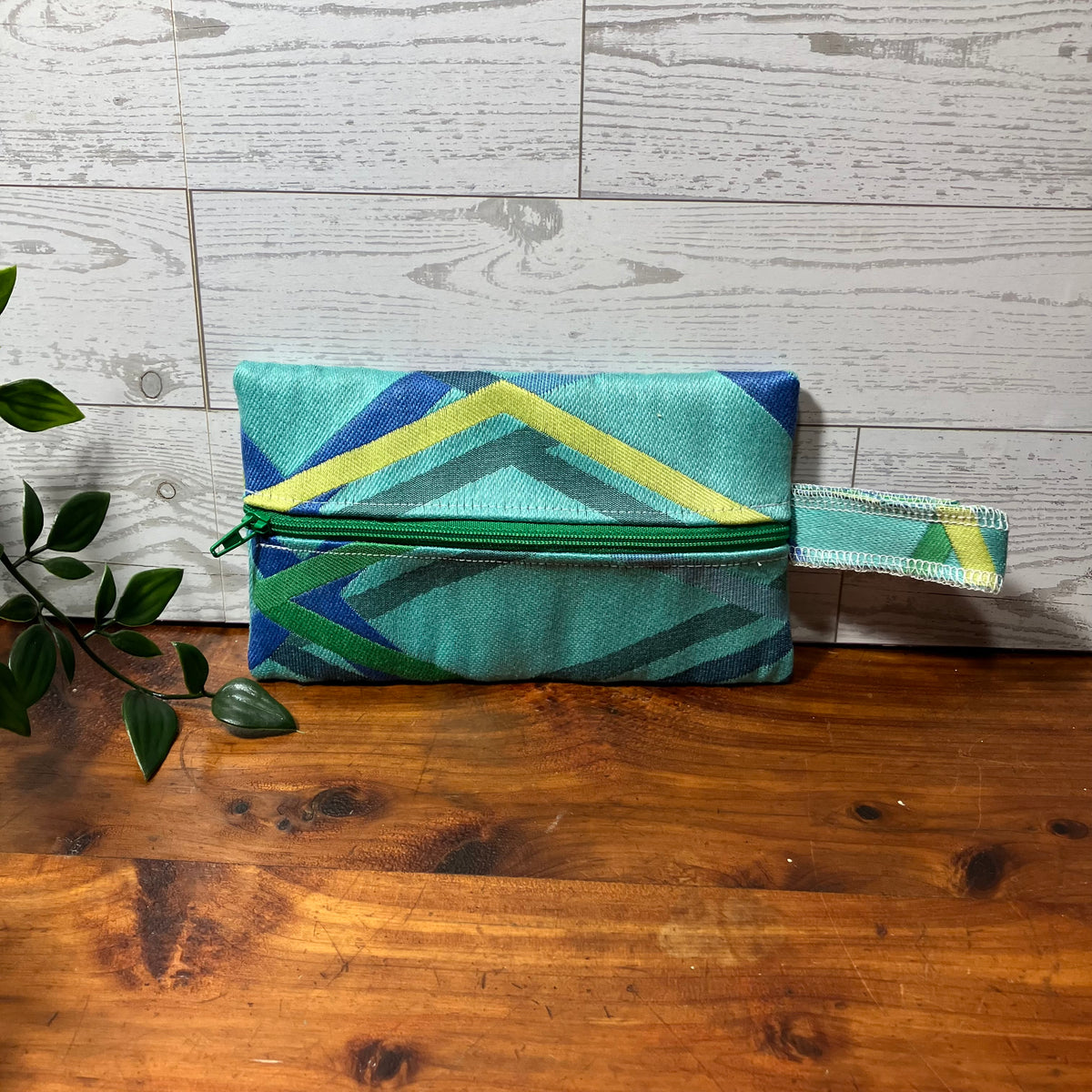 A fabric zippered pouch with a geometric pattern in teal, green, blue, and yellow, resting on a wooden surface against a white wood paneled wall.