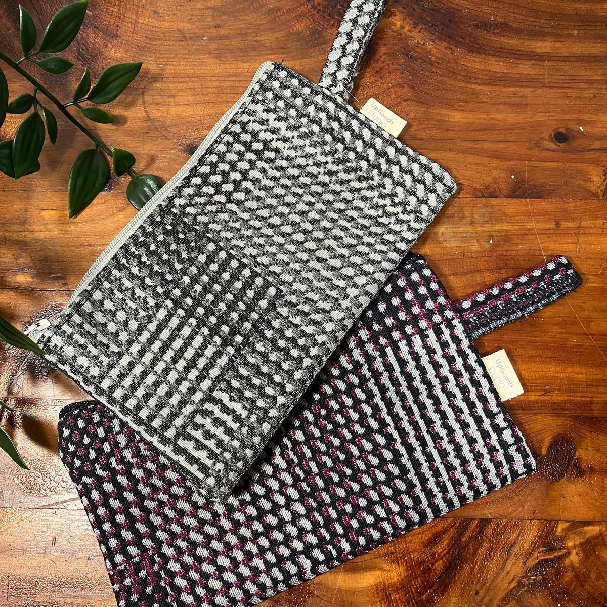 Two woven fabric pouches with top handles are arranged on a wooden tabletop. The upper pouch is black and white woven, and the lower pouch is black, white, and pink woven.