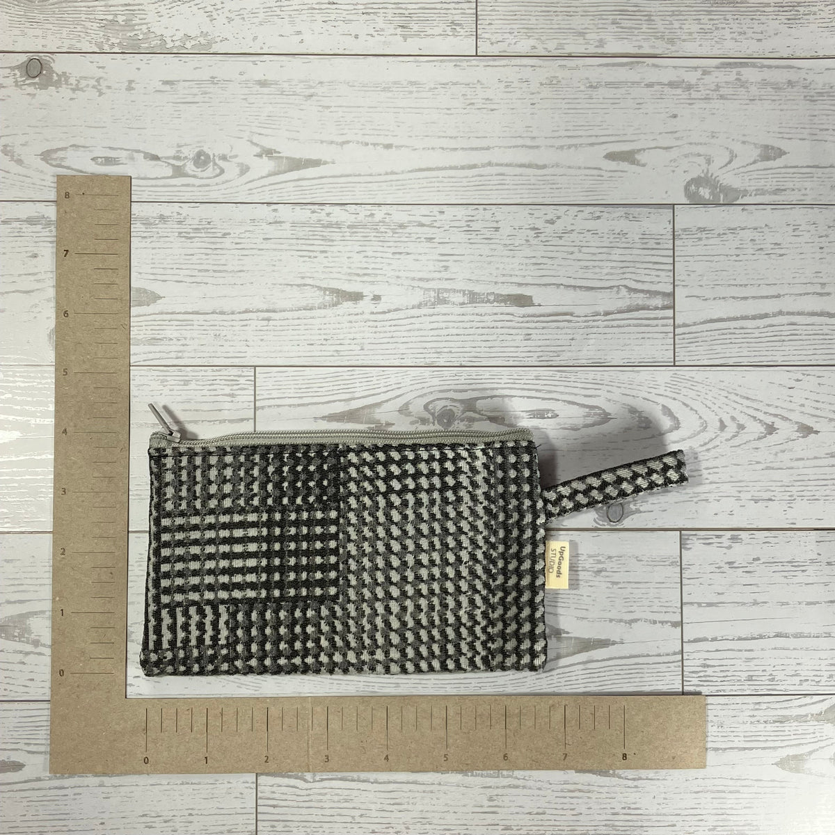 A woven black and white patterned clutch bag with a top handle, shown on a light gray wood plank background with cardboard rulers for scale.