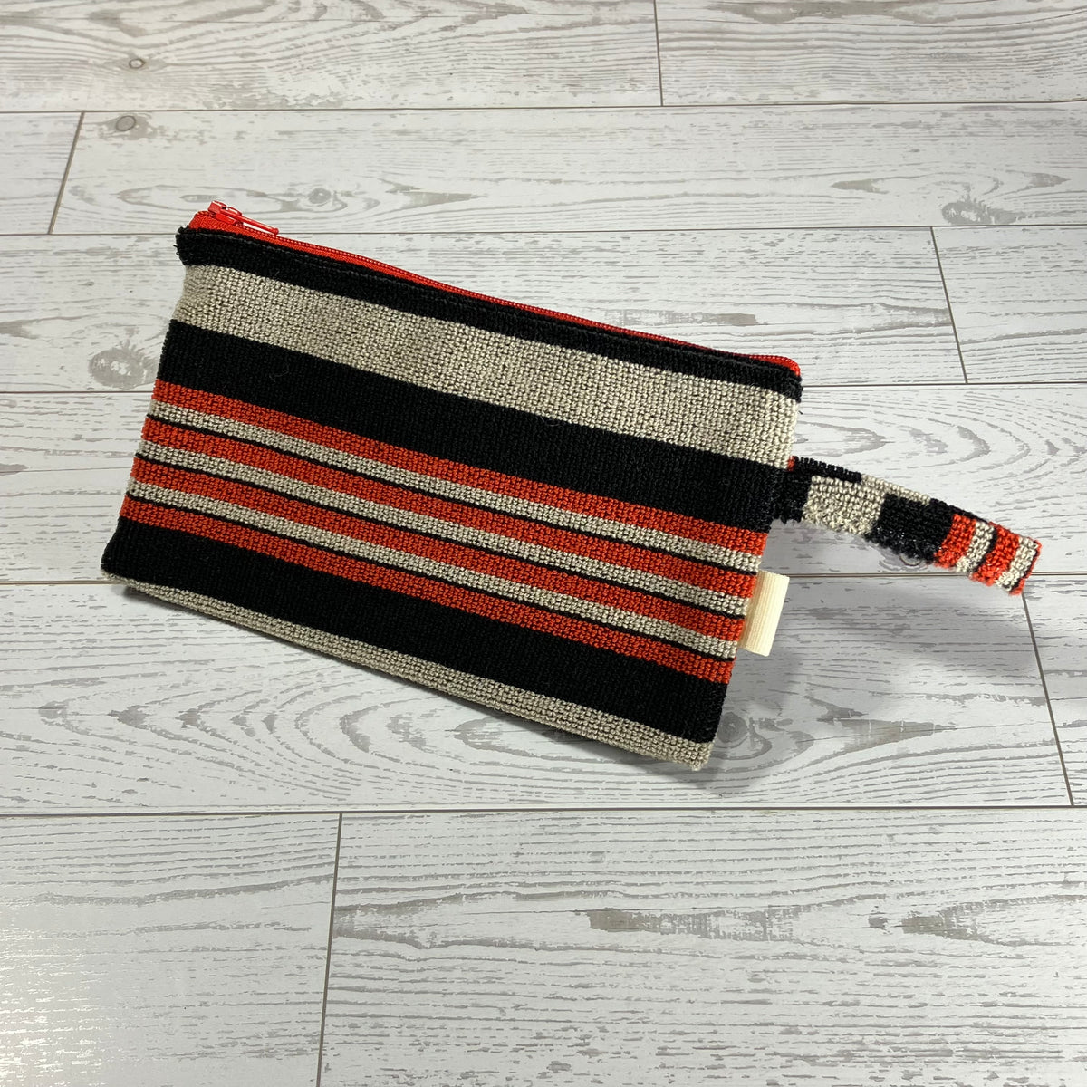 A woven zippered pouch with horizontal stripes in black, beige, and red, featuring a matching woven wristlet strap, placed on a light gray wood plank floor.