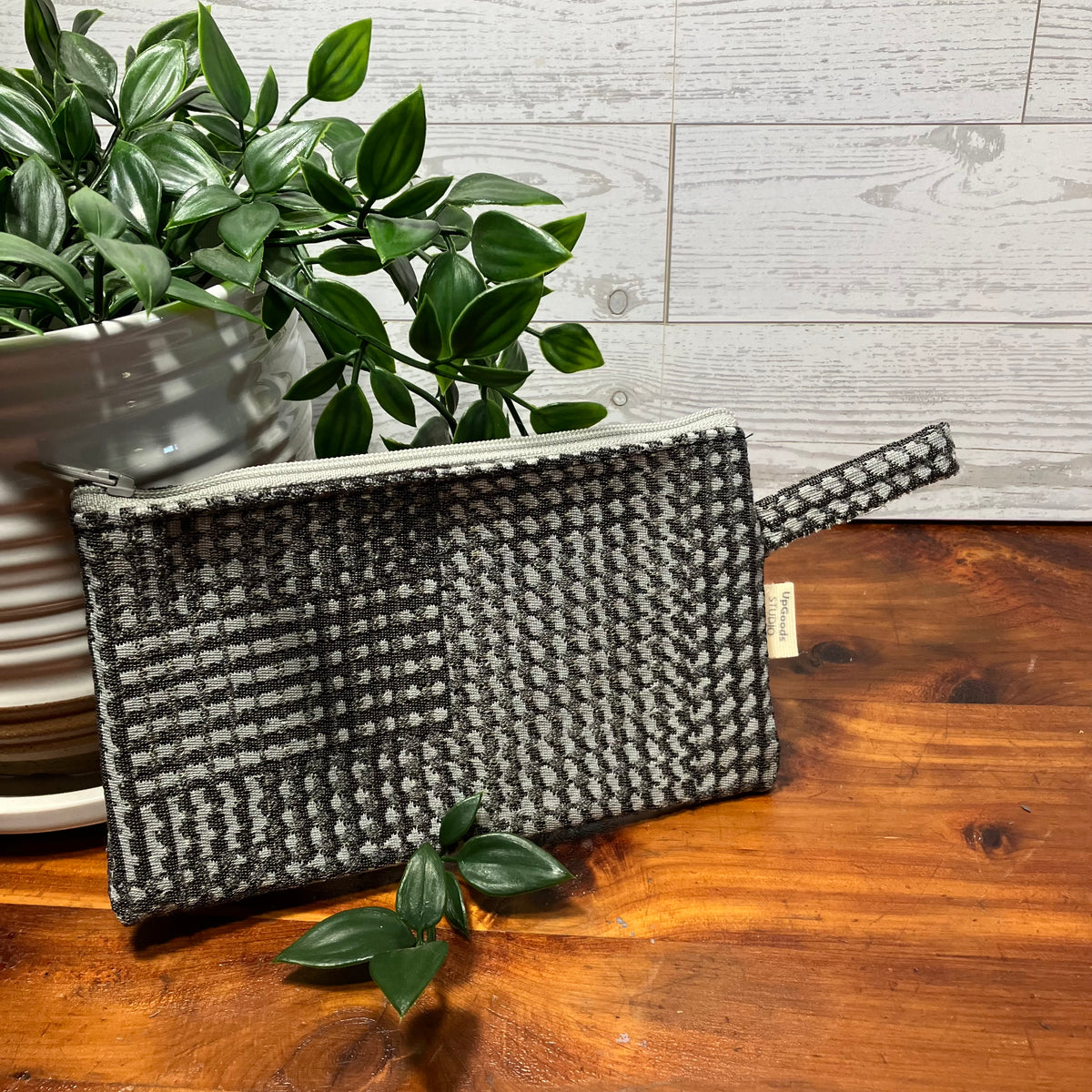 A gray and white woven houndstooth pouch with a top zipper and wrist strap, resting on a wooden tabletop next to a potted green plant.