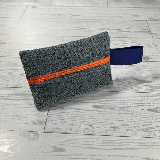 A dark gray textured fabric pouch with a bright orange zipper and a navy blue strap, placed on a light gray wood plank floor.