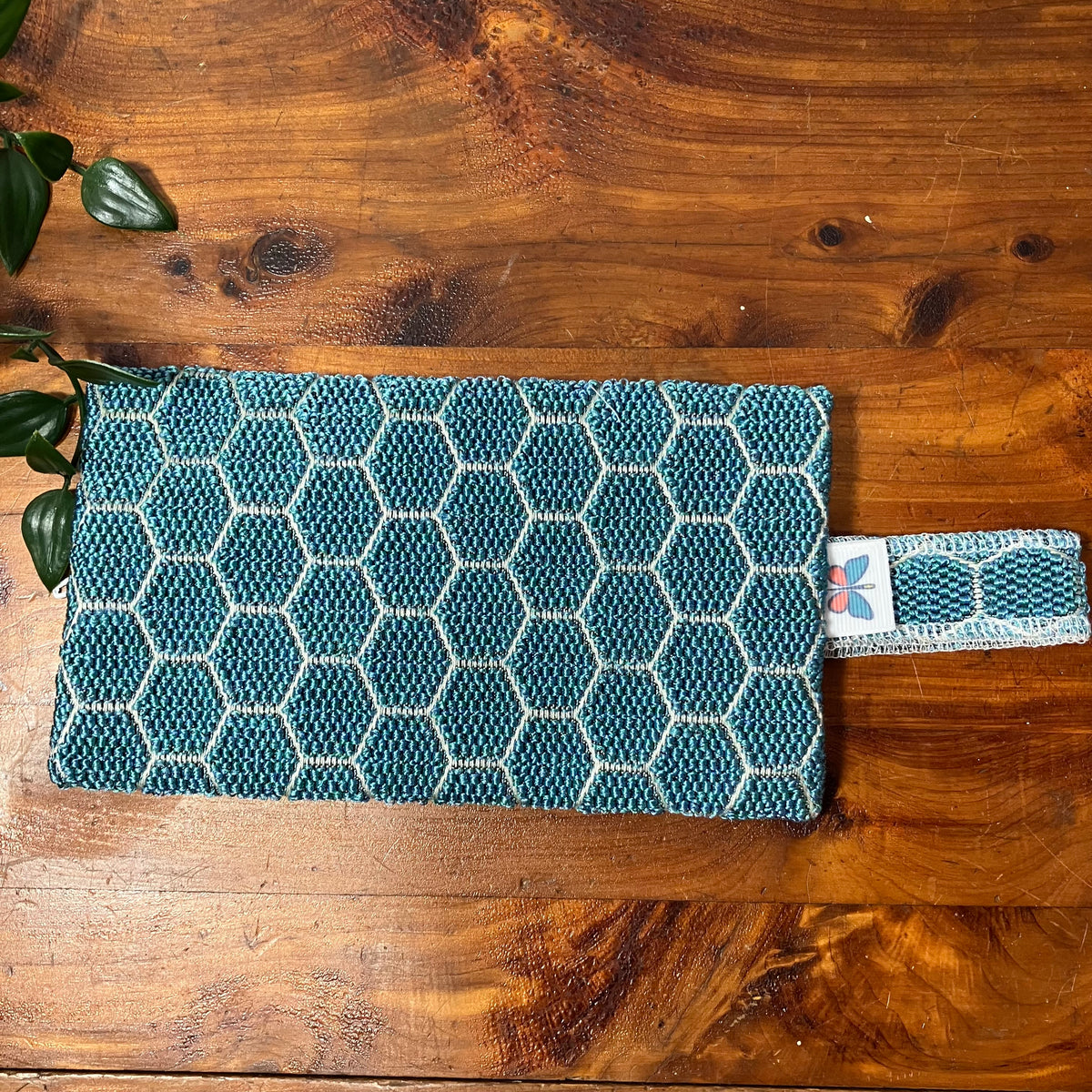 A rectangular woven clutch or pouch with a blue and white geometric honeycomb pattern, resting on a rustic wooden table with a small green leafy plant in the corner.