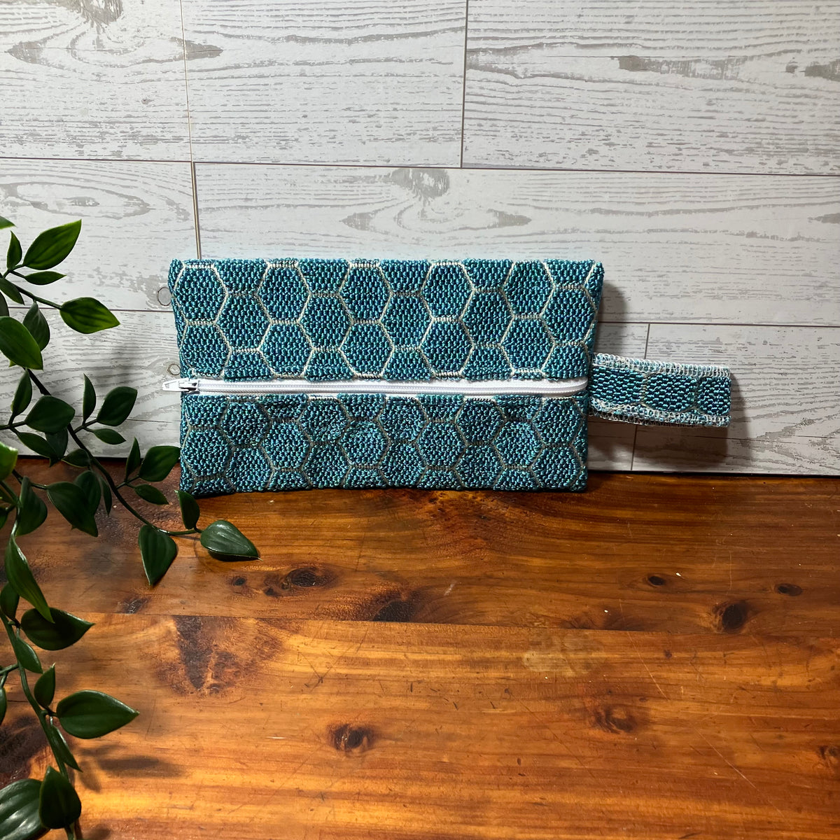 A teal and white patterned clutch sized pouch bag with a white zipper closure, resting on a wooden surface against a white wood-paneled wall, with a green plant branch entering from the left.