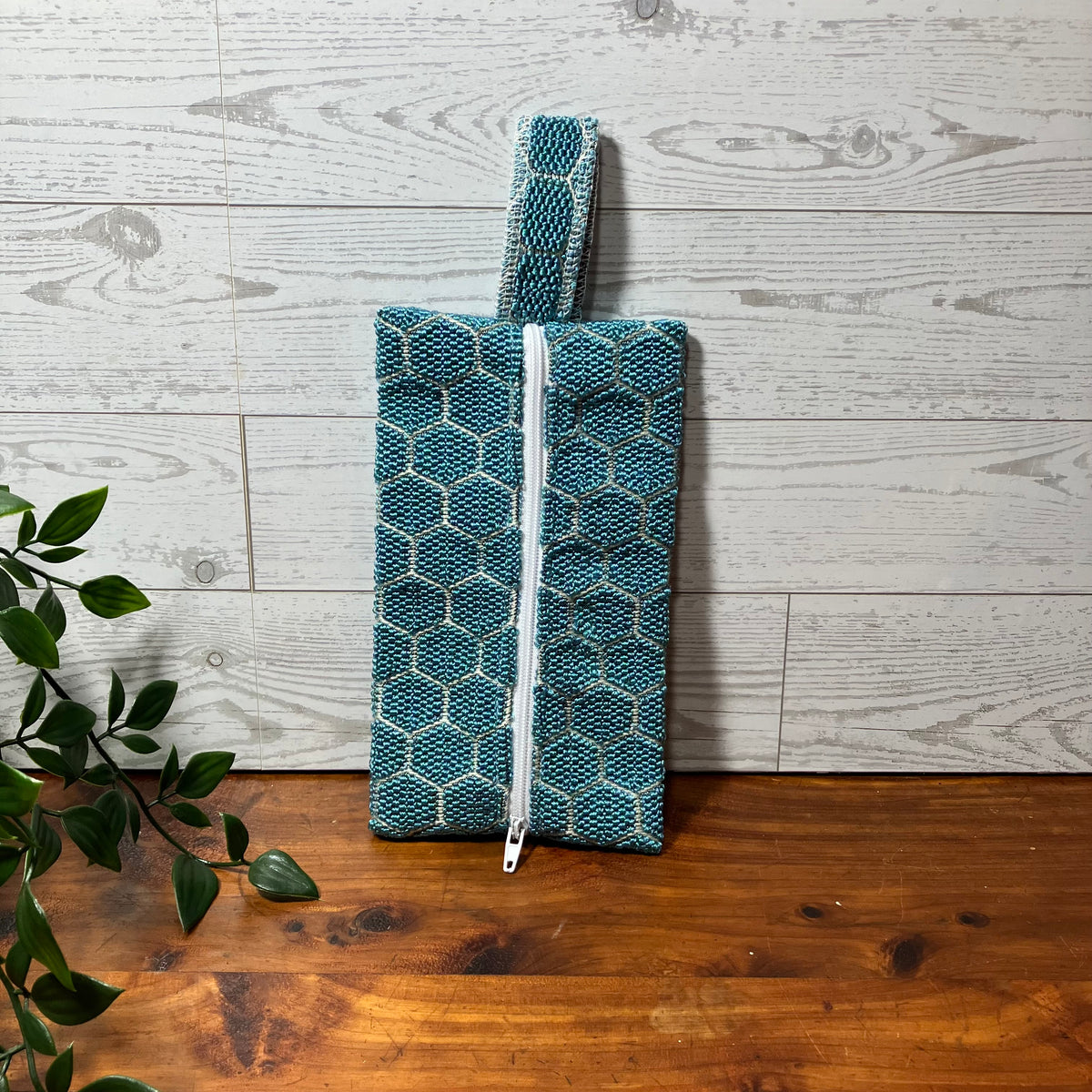 A teal and gray patterned fabric pouch with a white zipper and matching fabric loop handle, standing on a wooden surface against a whitewashed wood plank wall with green leaves entering from the left.