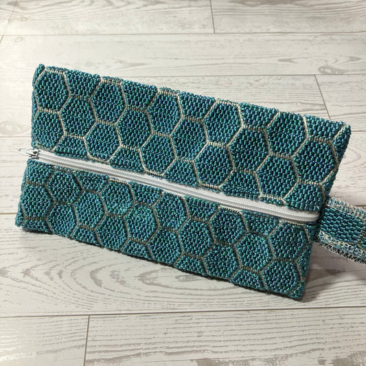 A teal and silver clutch sized pouch with a geometric honeycomb pattern, resting on a light gray wood plank floor.