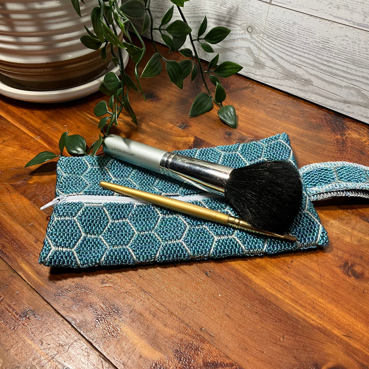 A teal and white patterned woven makeup pouch with a zipper closure, holding two makeup brushes, placed on a wooden tabletop with a potted plant in the background.