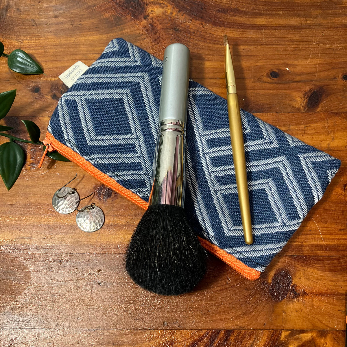 A geometric patterned zippered pouch with an orange zipper with a makeup brush resting on it, alongside earrings on a wooden tabletop.