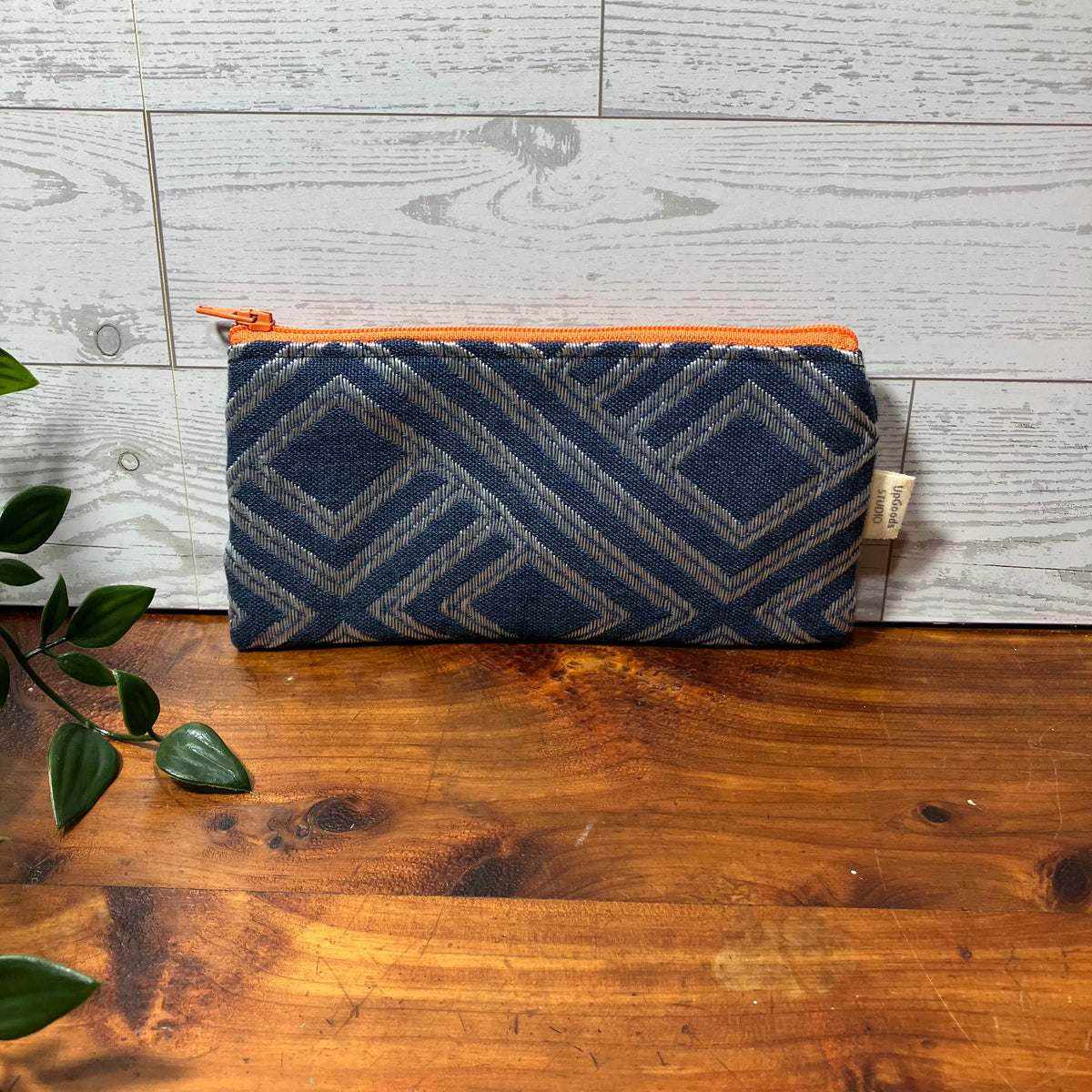 A zippered pouch with a navy blue and gray geometric patterned fabric body and a bright orange zipper, resting on a wooden tabletop against a light gray wood plank wall.
