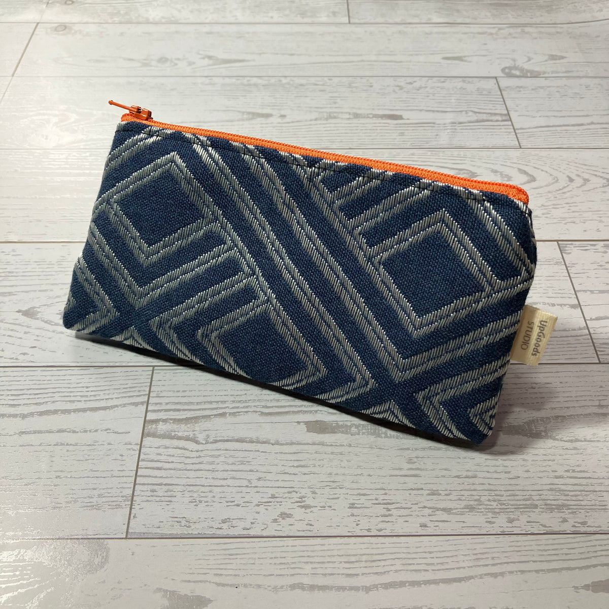 A zippered pouch with a navy blue and gray geometric patterned fabric, featuring a bright orange zipper and pull, resting on a light gray wood plank floor.
