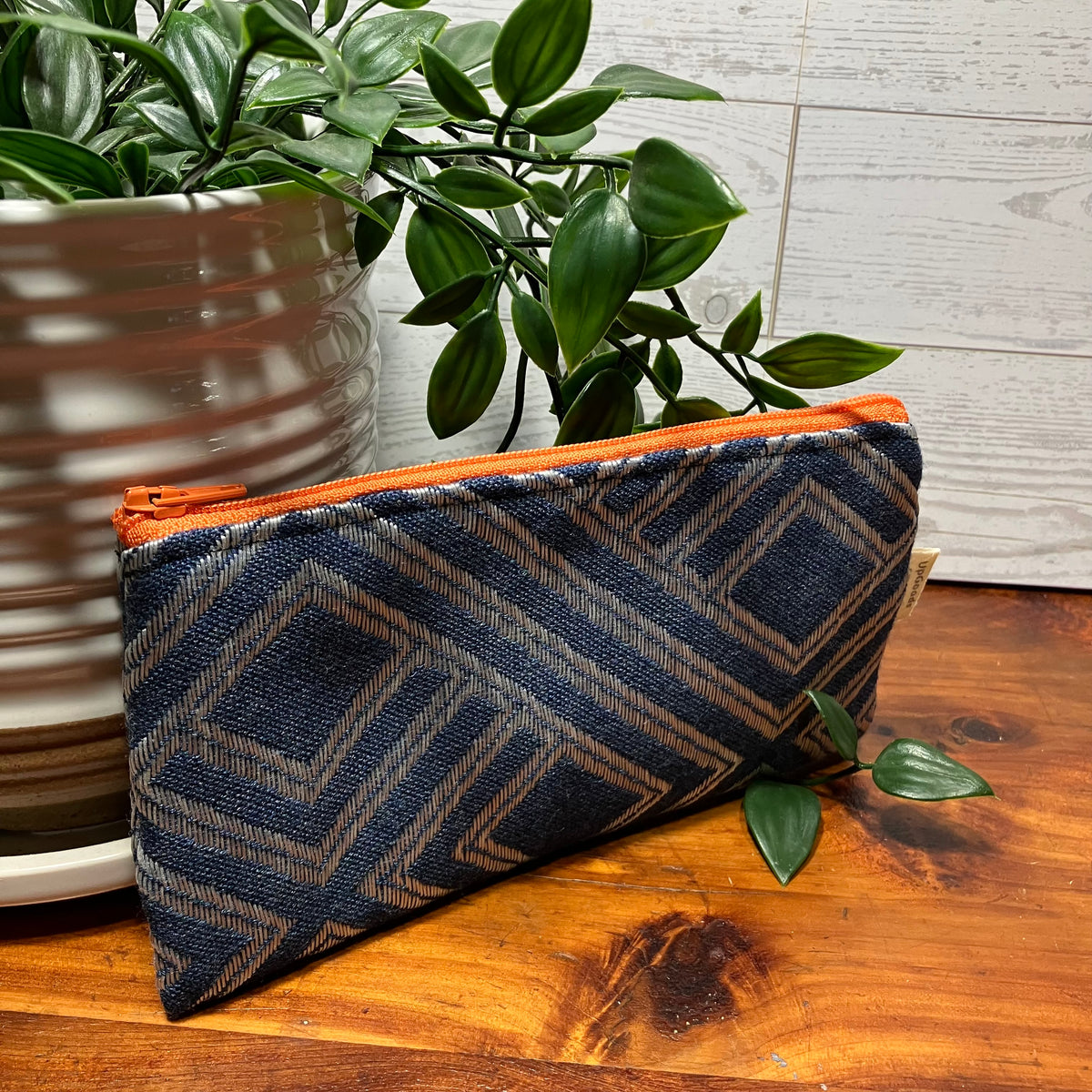 A zippered pouch with a navy and beige geometric patterned fabric, featuring an orange zipper, displayed on a wooden tabletop next to a potted green plant.