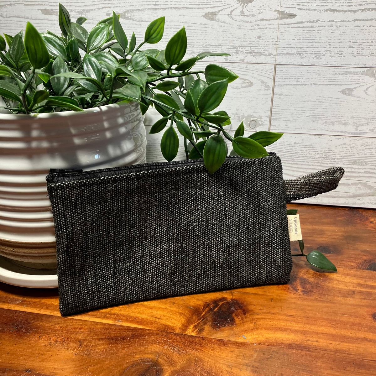 A black woven zip pouch with a matching wrist strap, placed on a wooden tabletop next to a potted green plant against a light wood plank wall.