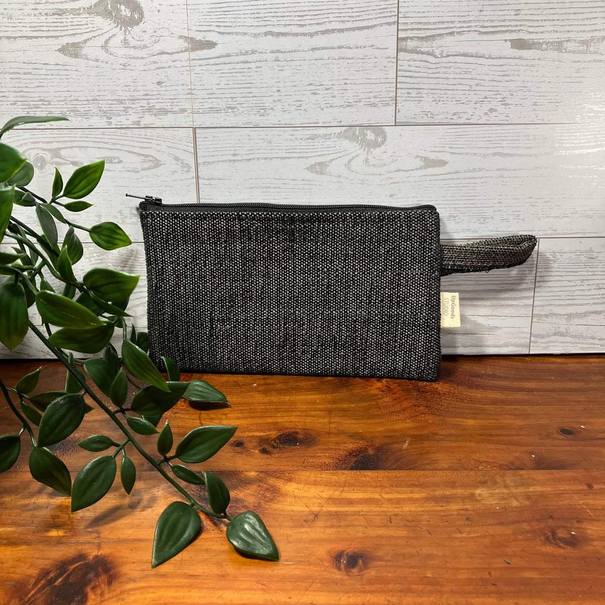 A dark gray woven zip pouch with a matching wrist strap, resting on a wooden surface against a light gray wood-paneled wall, with a green plant branch in the foreground.