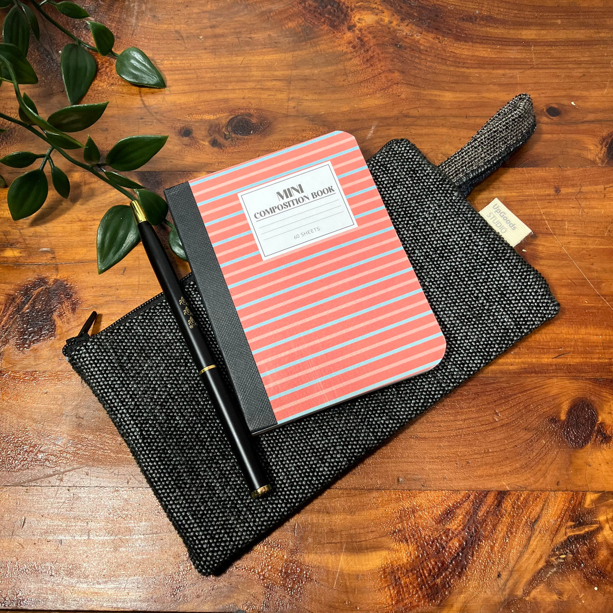 A black woven zip pouch with a handle, styled on a wooden tabletop with a coral and gray striped notebook and a black pen.