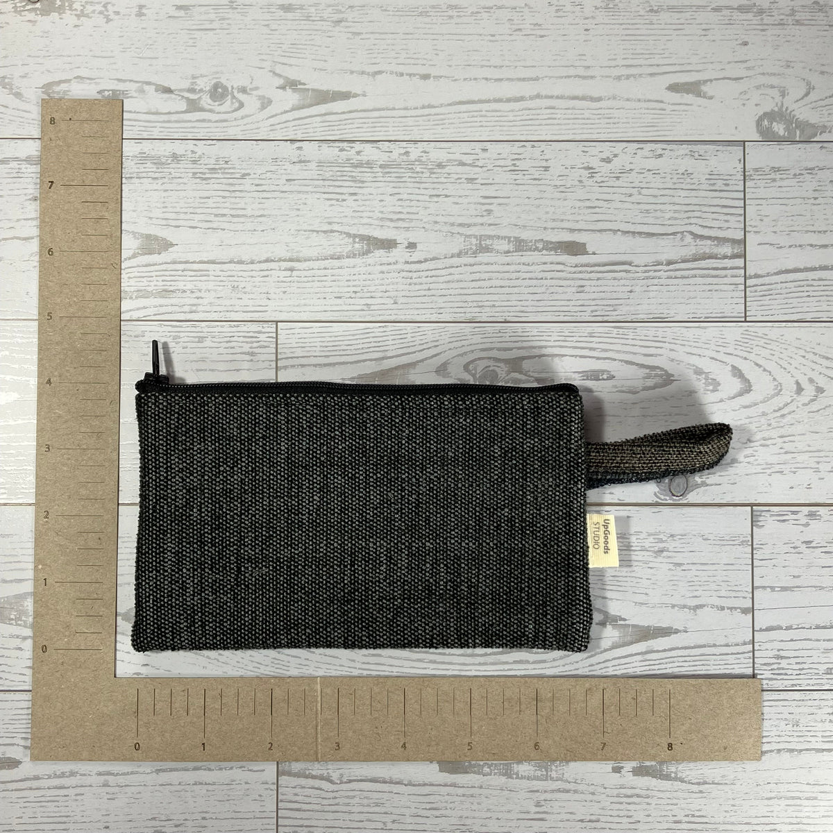 A black woven fabric zippered pouch with a matching loop handle, shown on a light gray wood-plank floor with cardboard rulers for scale.