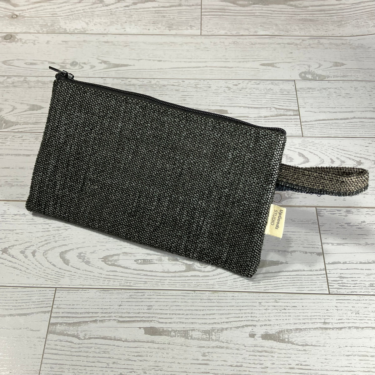 A dark gray/black woven zippered pouch with a matching wristlet strap and a small beige tag, placed on a light gray wood plank floor.