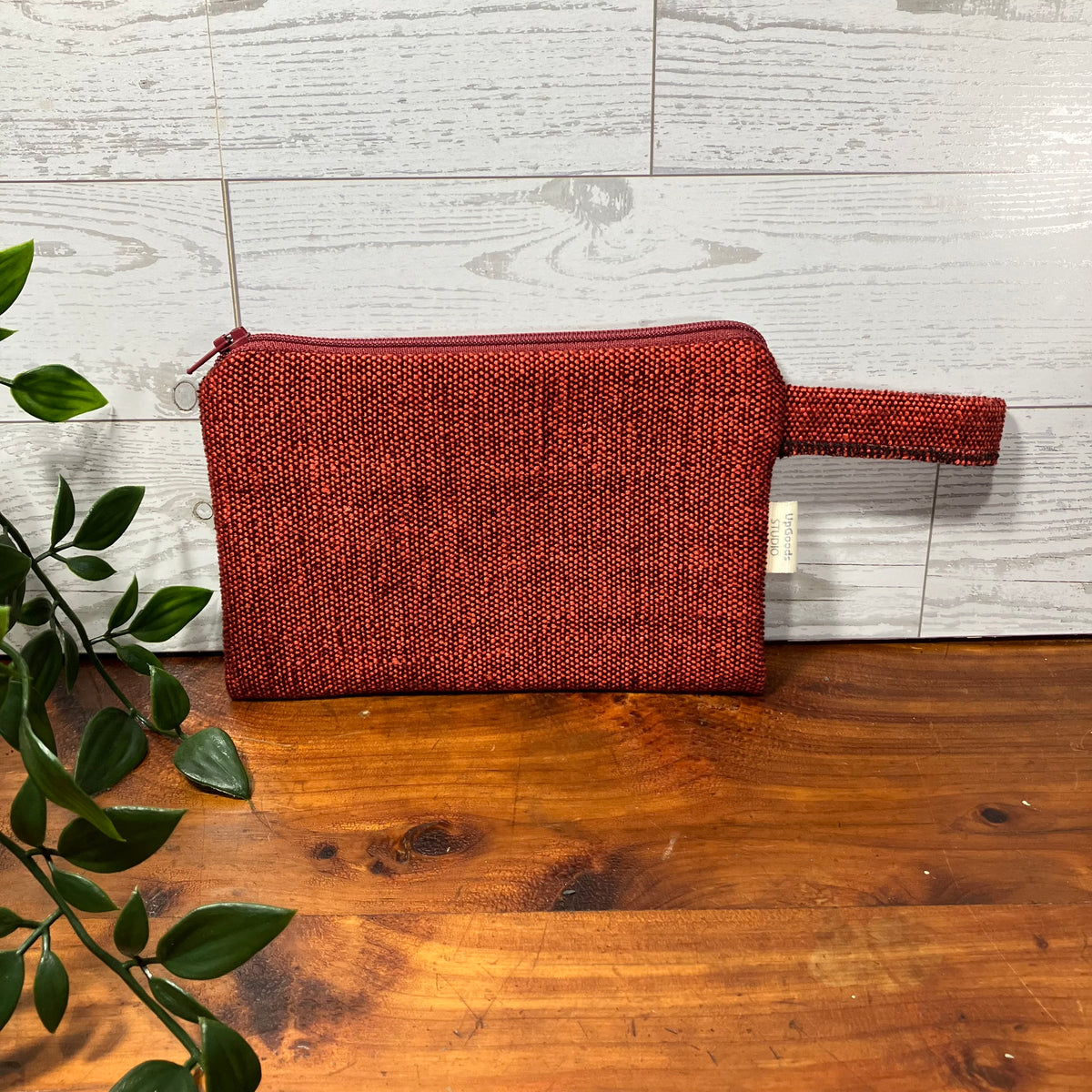 A textured red woven cosmetic pouch with a top zipper and integrated wrist strap, resting on a wooden tabletop against a white wood plank wall, with green leaves entering from the left side.