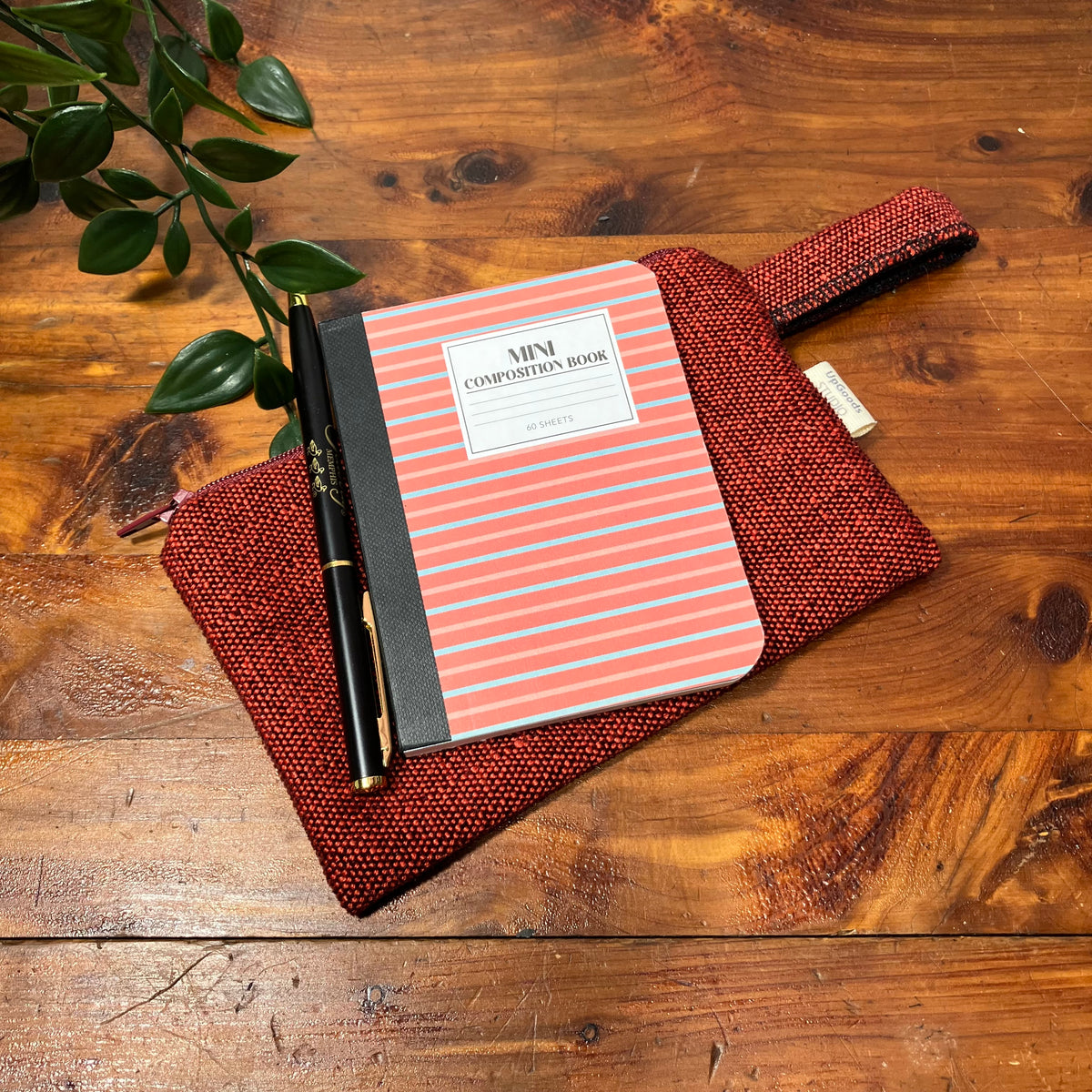 A woven red zippered pouch with a matching loop strap, holding a striped notebook and a black pen, arranged on a rustic wooden tabletop with a green plant leaf in the corner.