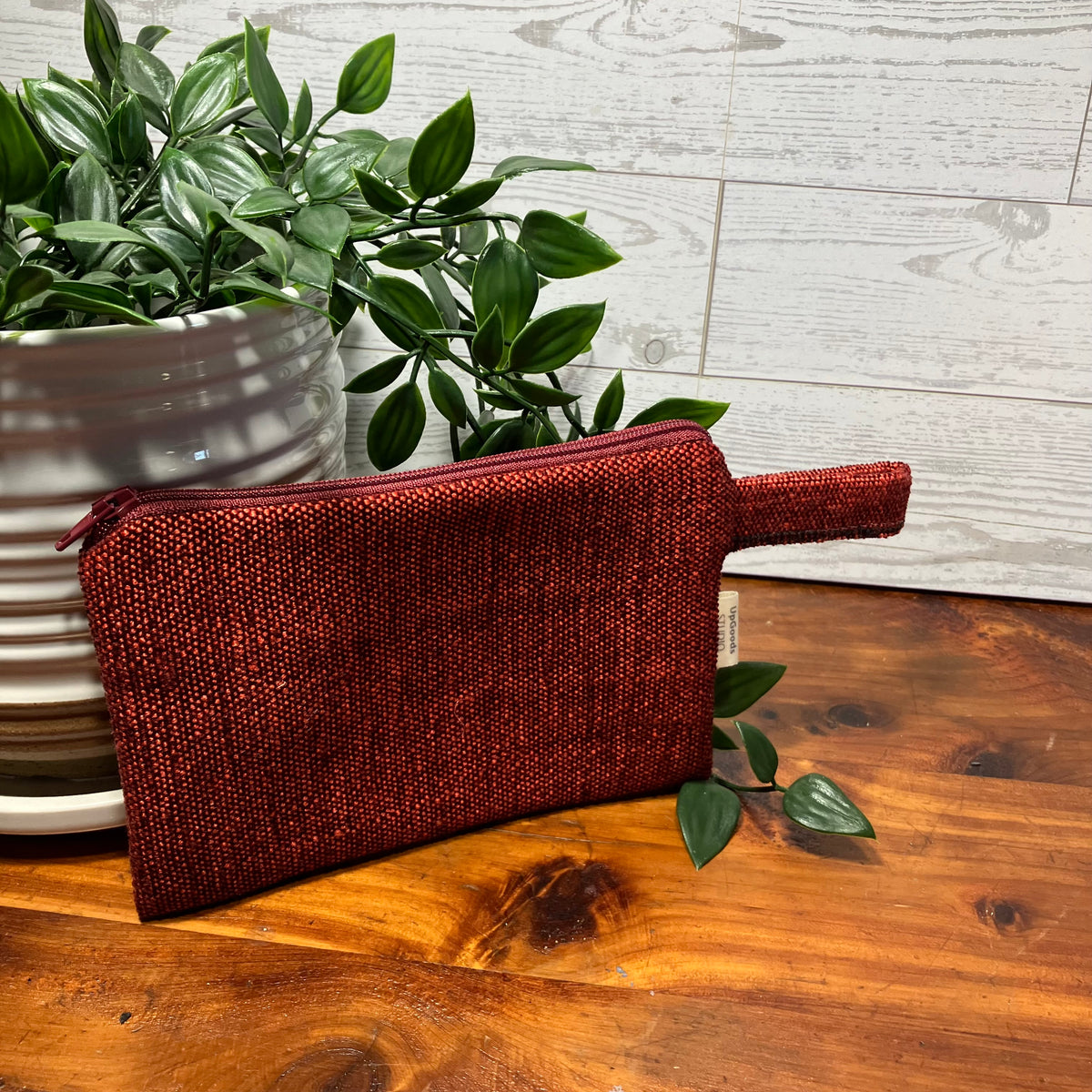A textured, metallic deep red zippered pouch with a handle, resting on a wooden tabletop next to a potted green plant.
