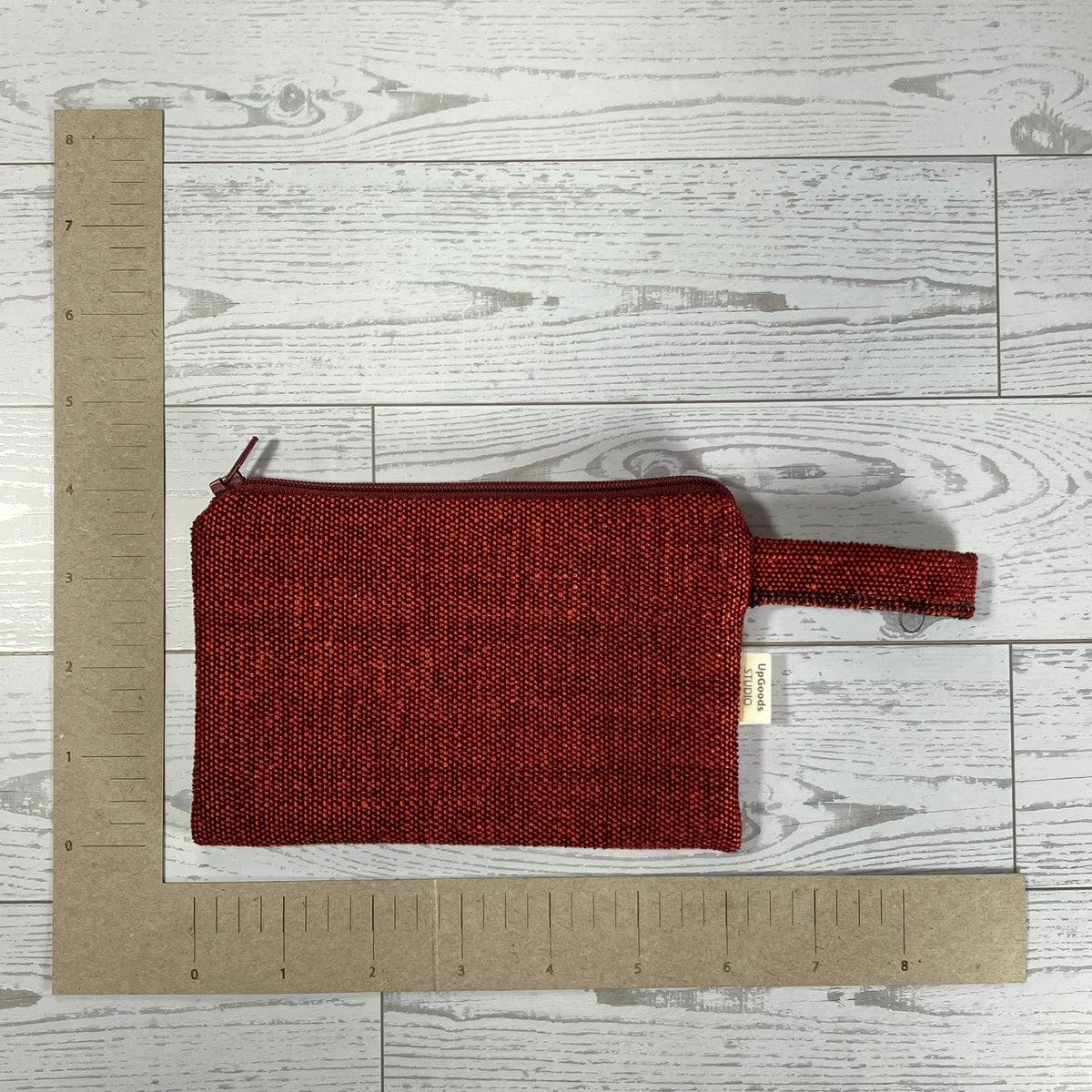 A small woven zip pouch in a deep rust red color with a matching loop handle, laid flat on a light gray wood plank surface. A cardboard ruler is positioned vertically and horizontally to show the product's dimensions.