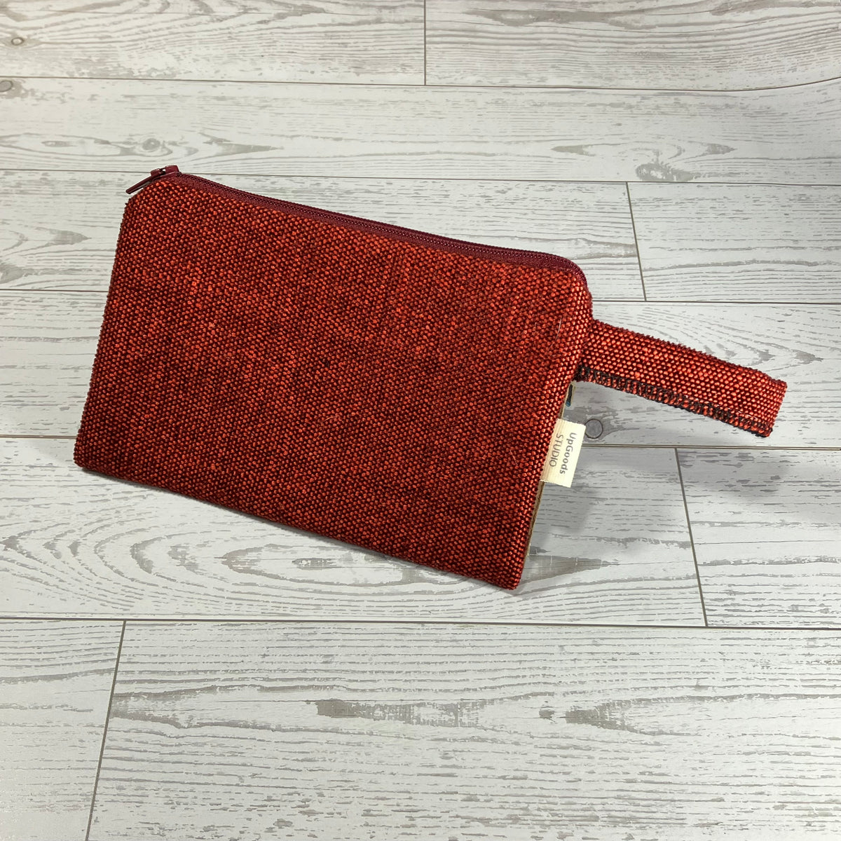 A textured red zippered pouch with a matching wristlet strap, resting on a light gray wood-plank floor. A small cream-colored fabric tag is attached to the side.