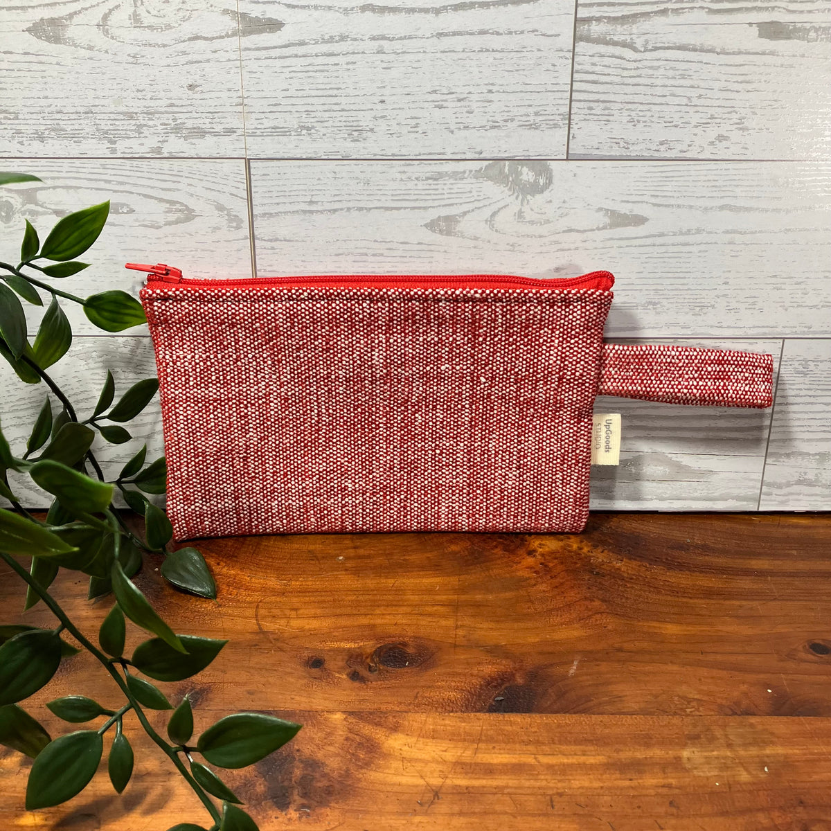 A woven red and white zippered pouch with a matching wrist strap, resting on a wooden surface against a light gray wood plank wall, with green leaves entering from the left.