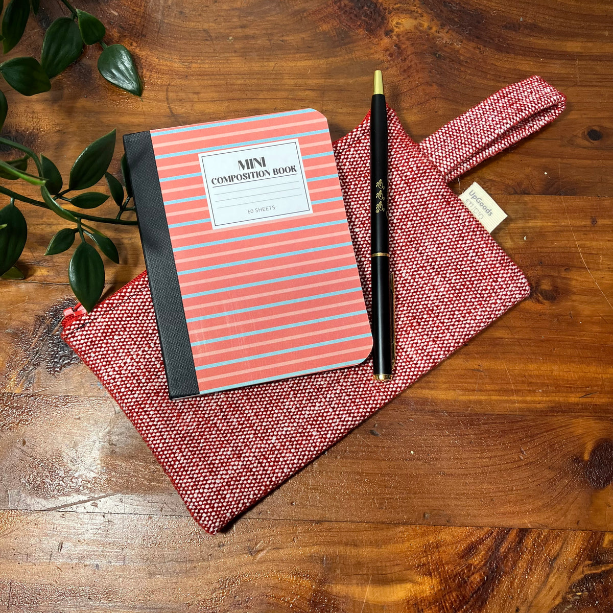 A red woven fabric pouch arranged with a mini composition notebook with coral and gray stripes, and a black pen on a wooden tabletop with green leaves in the corner.