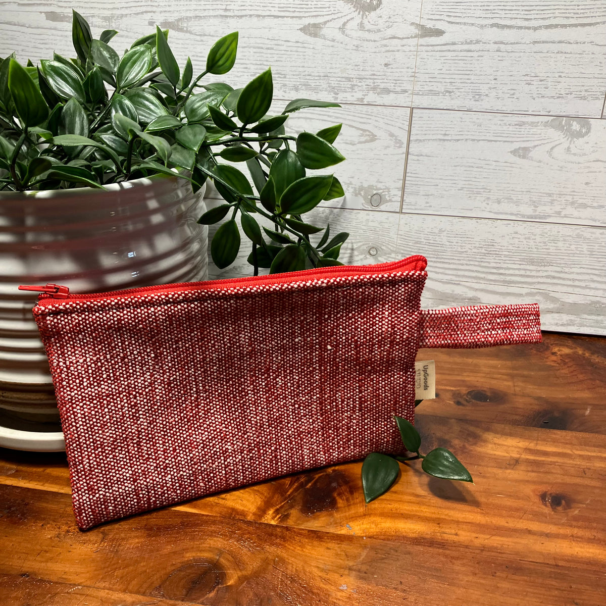 A textured red and silver woven zippered pouch with a small fabric tag, resting on a wooden table next to a potted green plant.