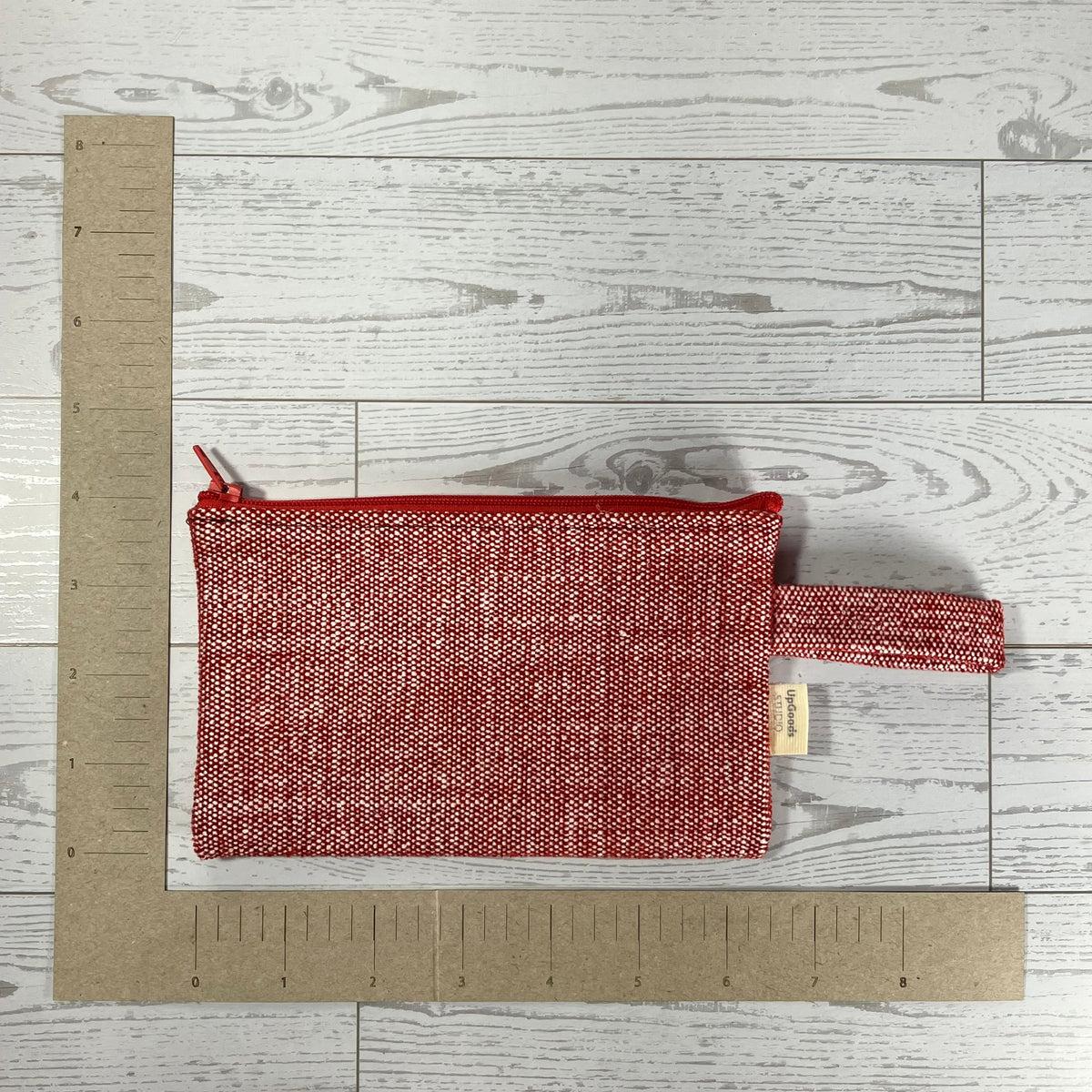 A small woven zippered pouch with a top handle in a red and white textured fabric, laid flat on a light gray wood plank surface next to cardboard rulers for scale.