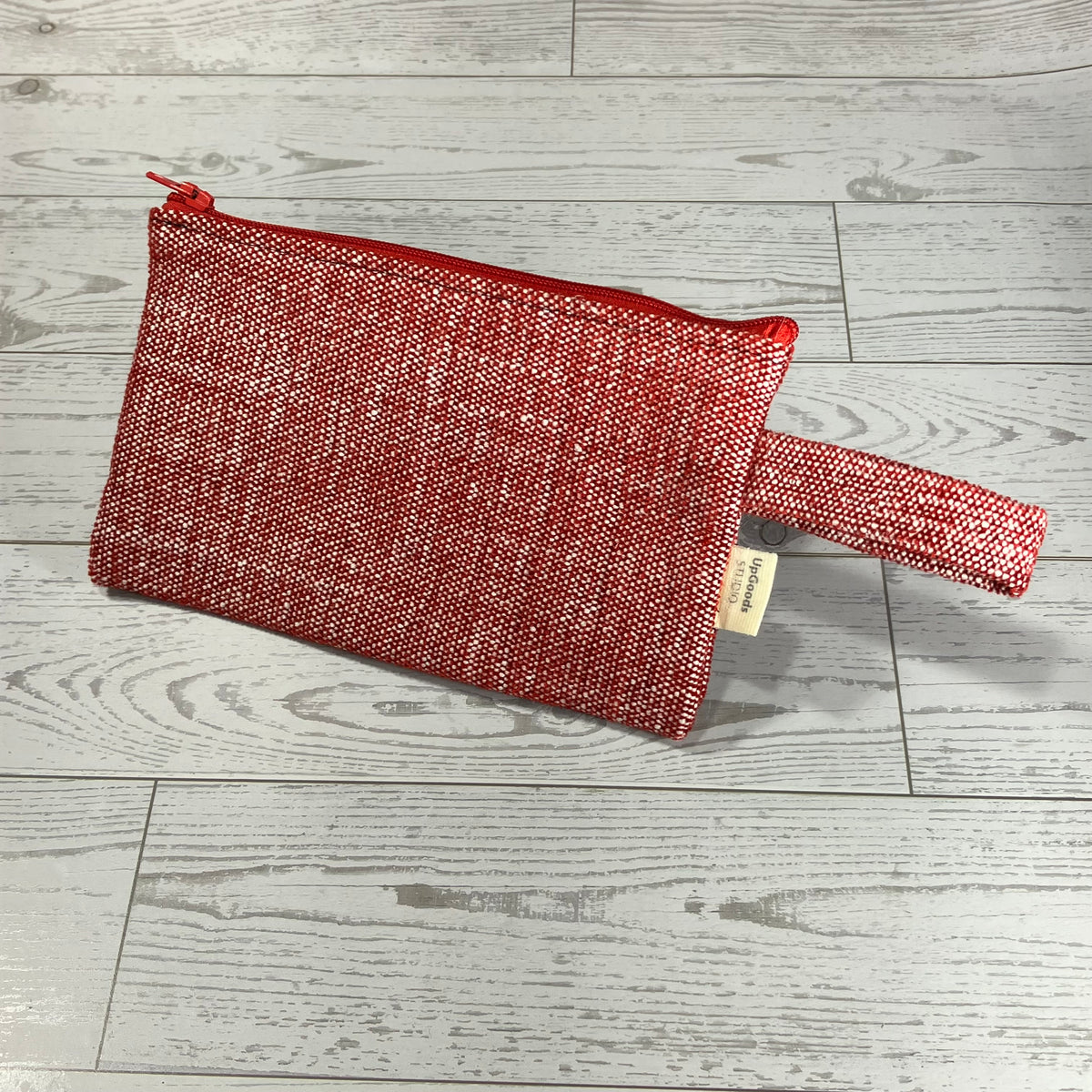 A red and white textured woven zip pouch with a matching wristlet strap, placed on a light gray wood plank floor.