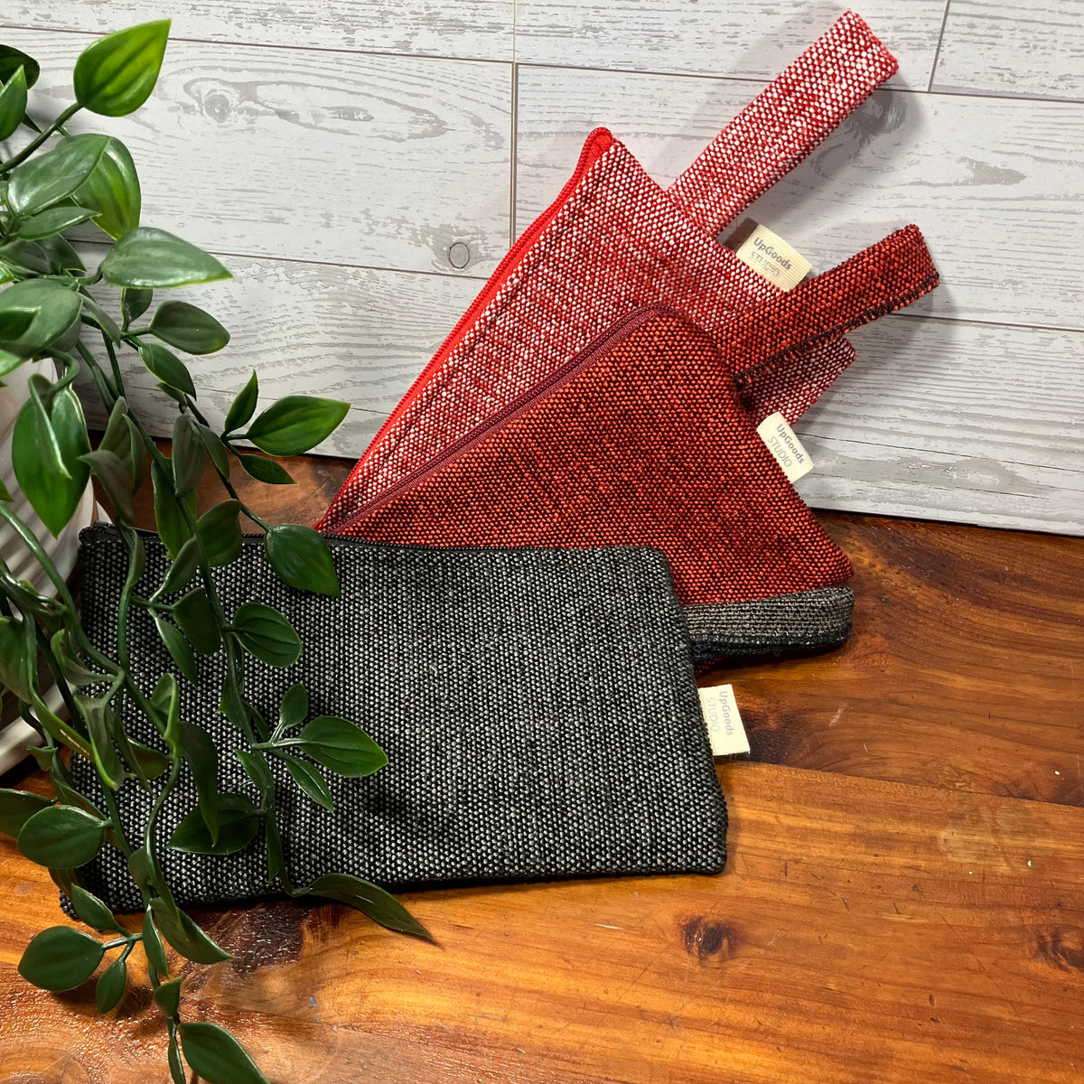 Three woven fabric pouches or small bags in black, red, and rust tones arranged on a wooden surface with a white wood plank backdrop and a green plant leaf in the foreground.