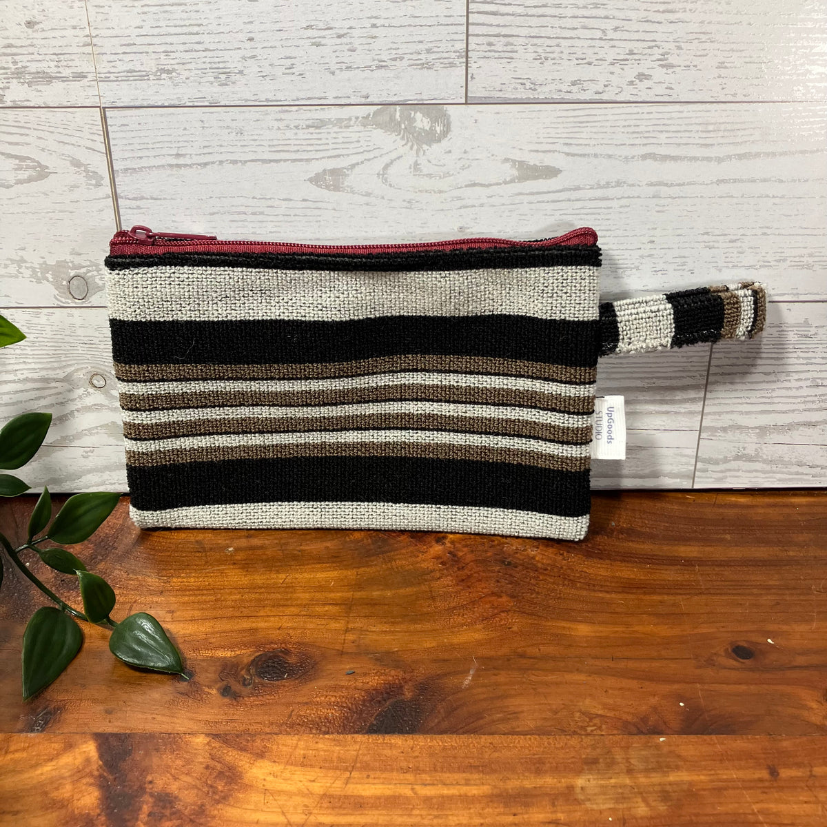 A woven zip pouch with horizontal black, brown, and beige stripes, featuring a dark red zipper and a matching striped wrist strap, displayed on a wooden surface against a light wood paneled background.