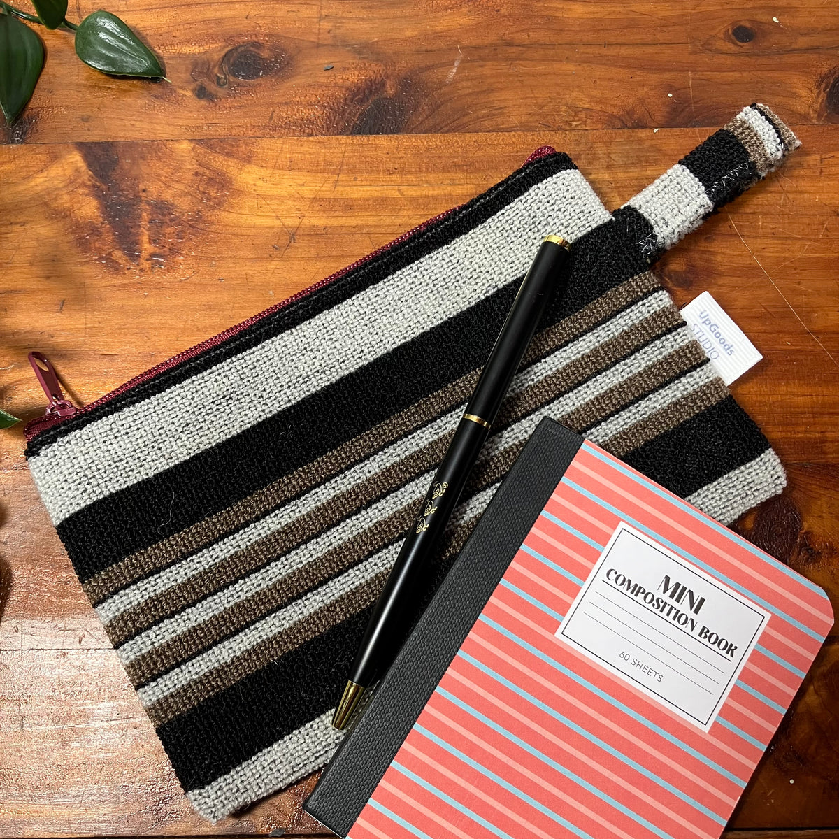 A woven zippered pouch with horizontal black, beige, and brown stripes, styled on a wooden tabletop alongside a black pen and a coral-pink composition notebook.
